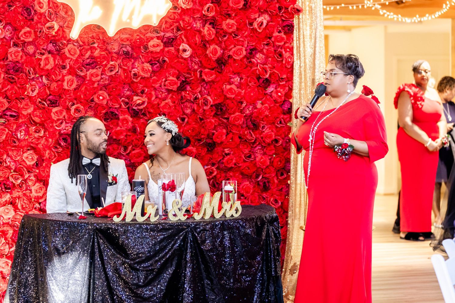 Wedding reception setup with sweetheart table against stunning red rose wall backdrop.