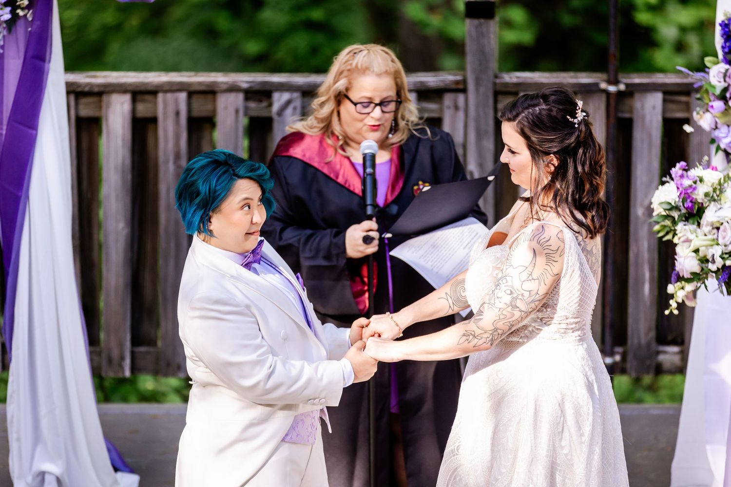 A heartfelt wedding ceremony takes place on a wooden deck decorated with purple draping and floral arrangements.