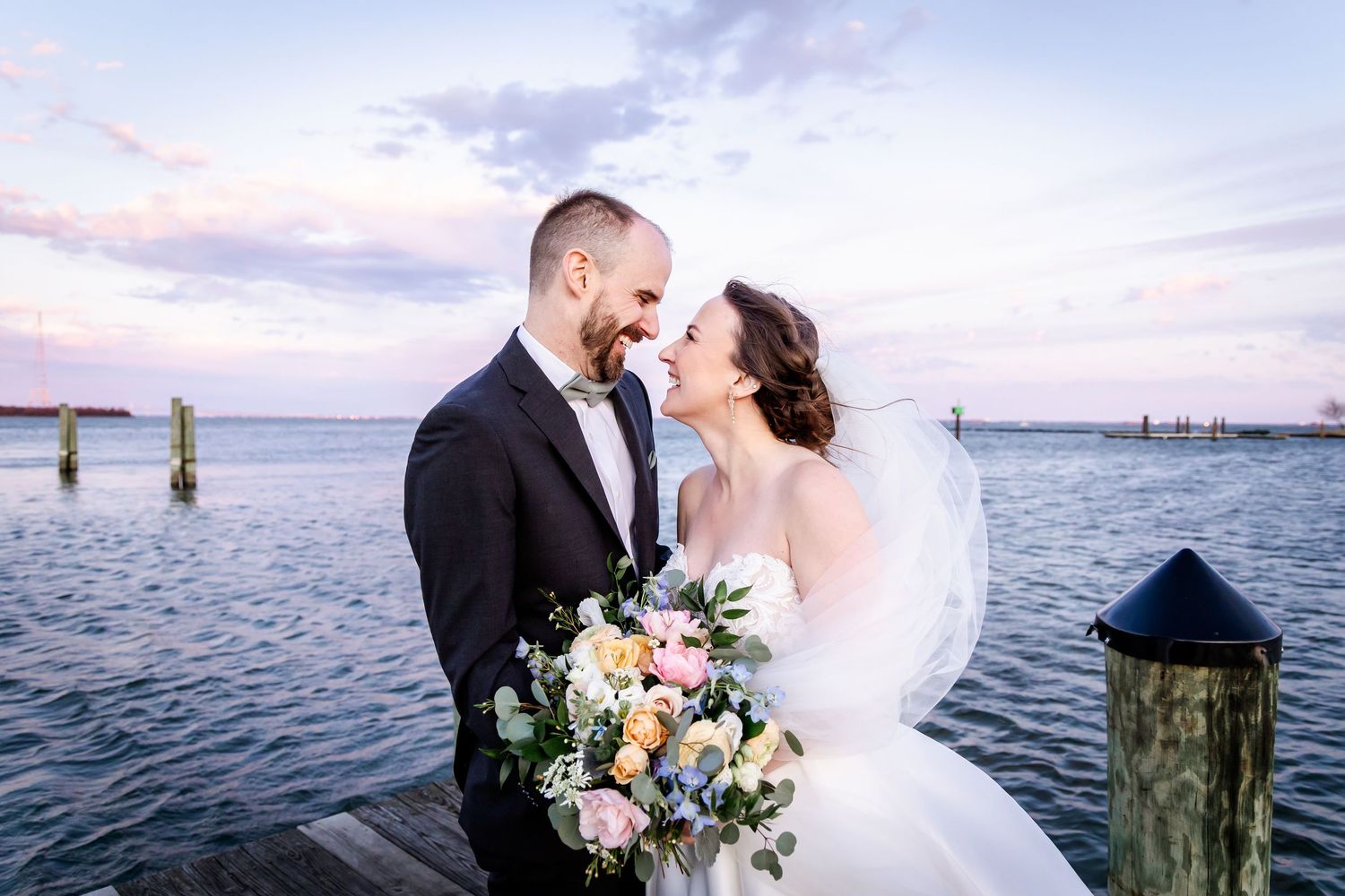 A romantic wedding couple stands on a dock at sunset holding a beautiful bouquet beside the calm waterfront.