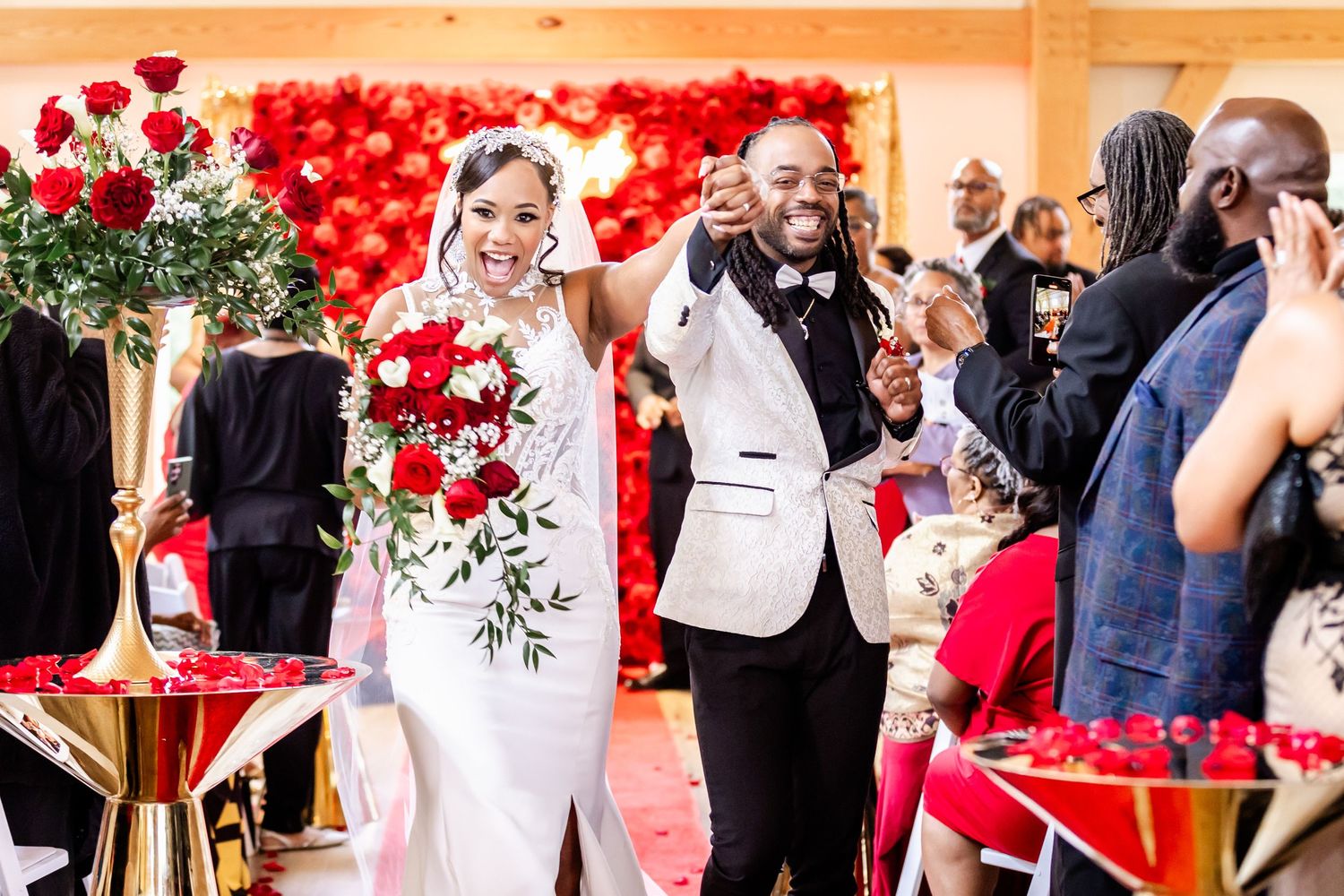 Newlyweds celebrating joyfully at their wedding reception with a red flower wall behind them.