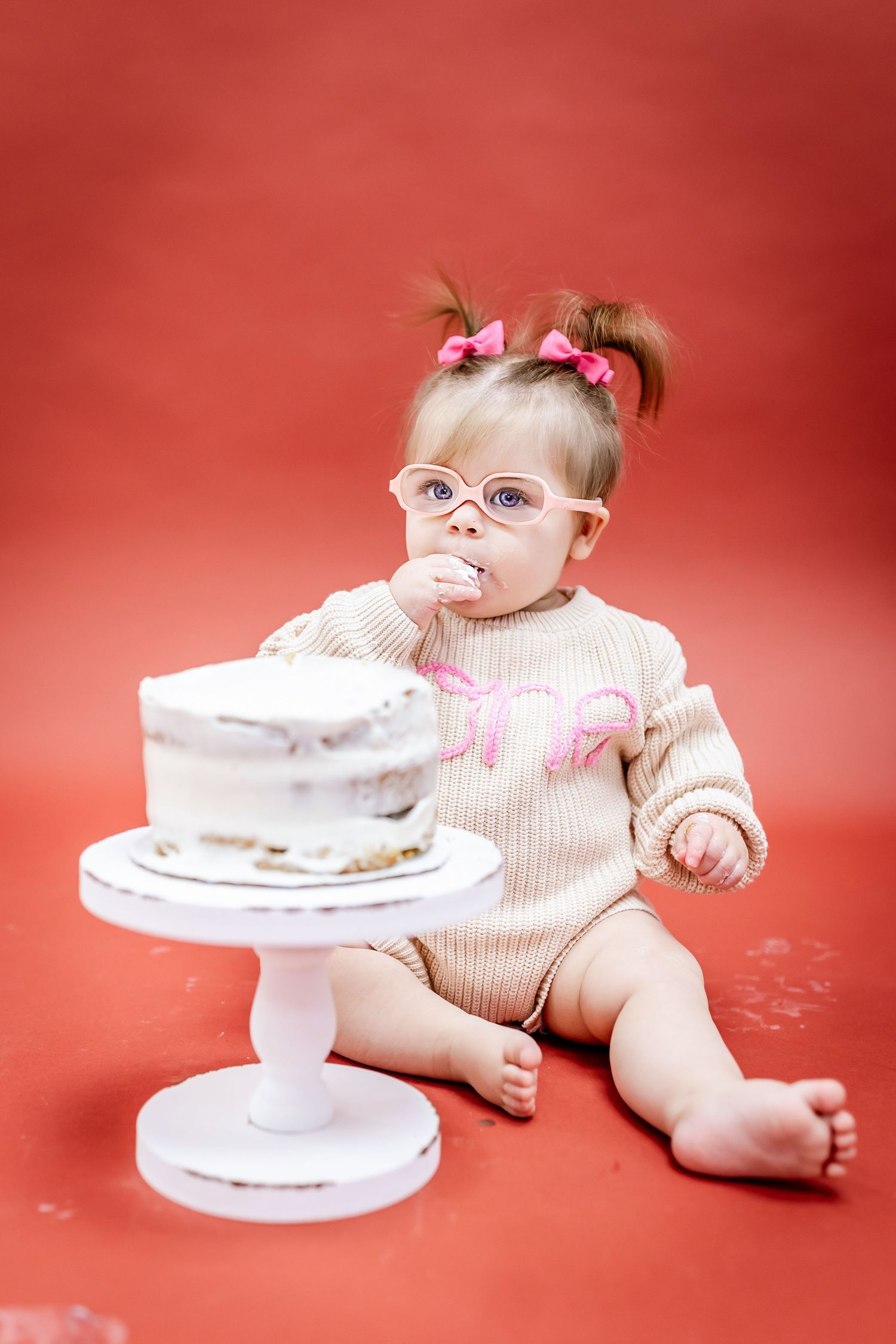 Baby in knit outfit and glasses looks at white cake on pedestal against red backdrop.