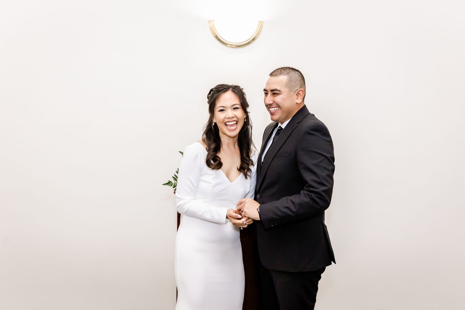 Happy newlyweds share a joyful moment together against a white wall with decorative lighting.