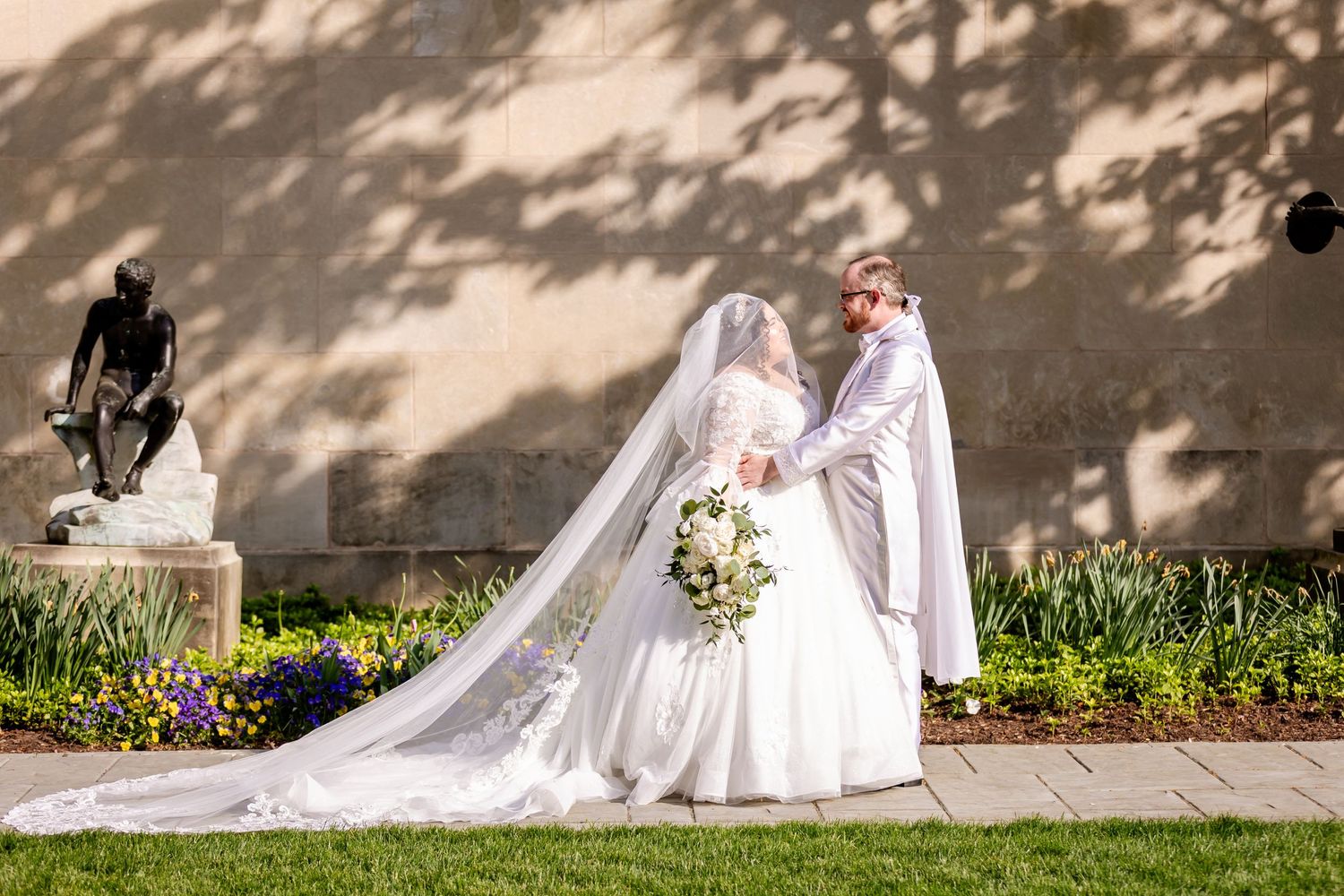 A bride and groom pose together on their wedding day against a sunlit wall with dappled shadows.