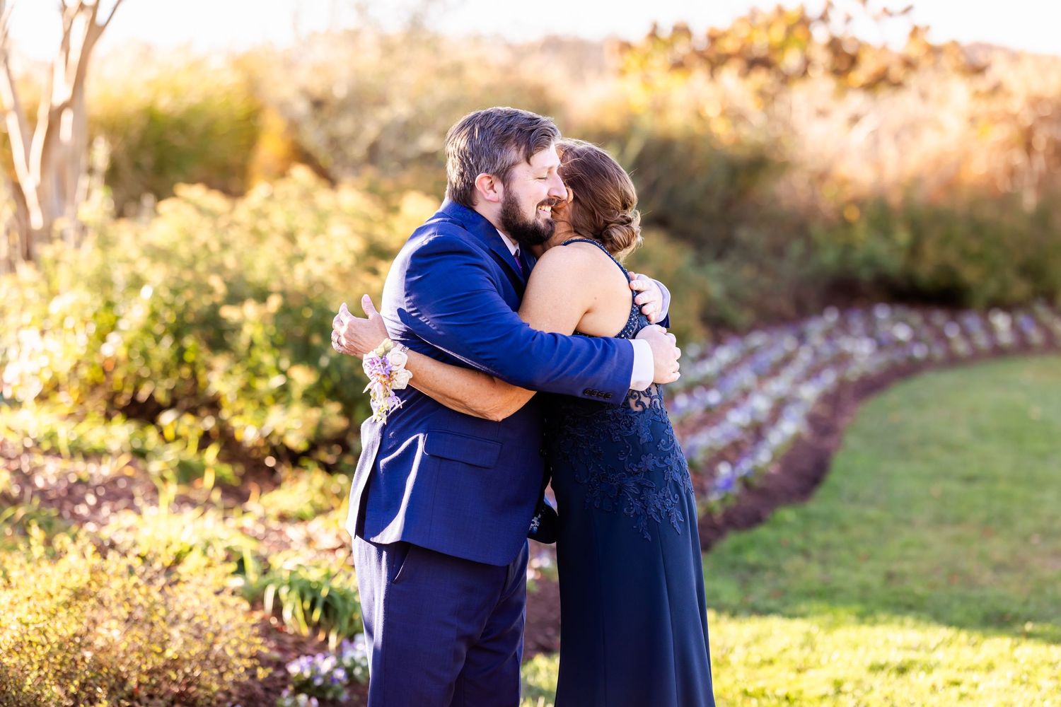 Couple sharing an intimate embrace outdoors at sunset in formal navy blue attire by a garden path.