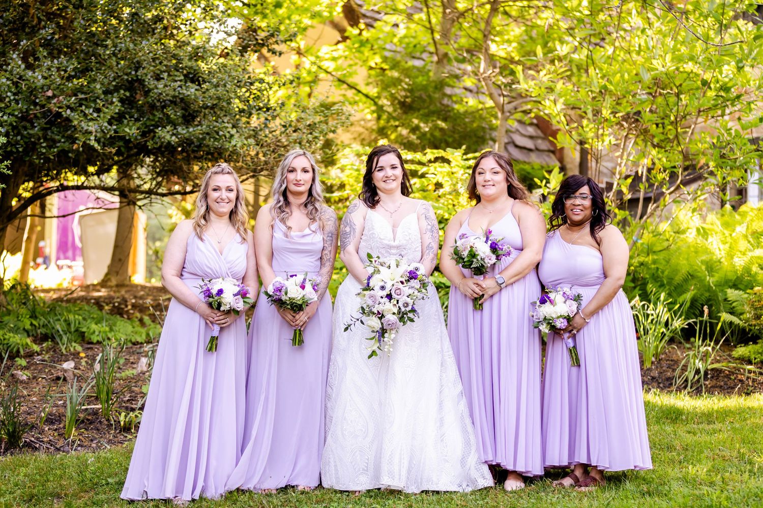Bridal party in lavender dresses posed together in sunny garden setting.