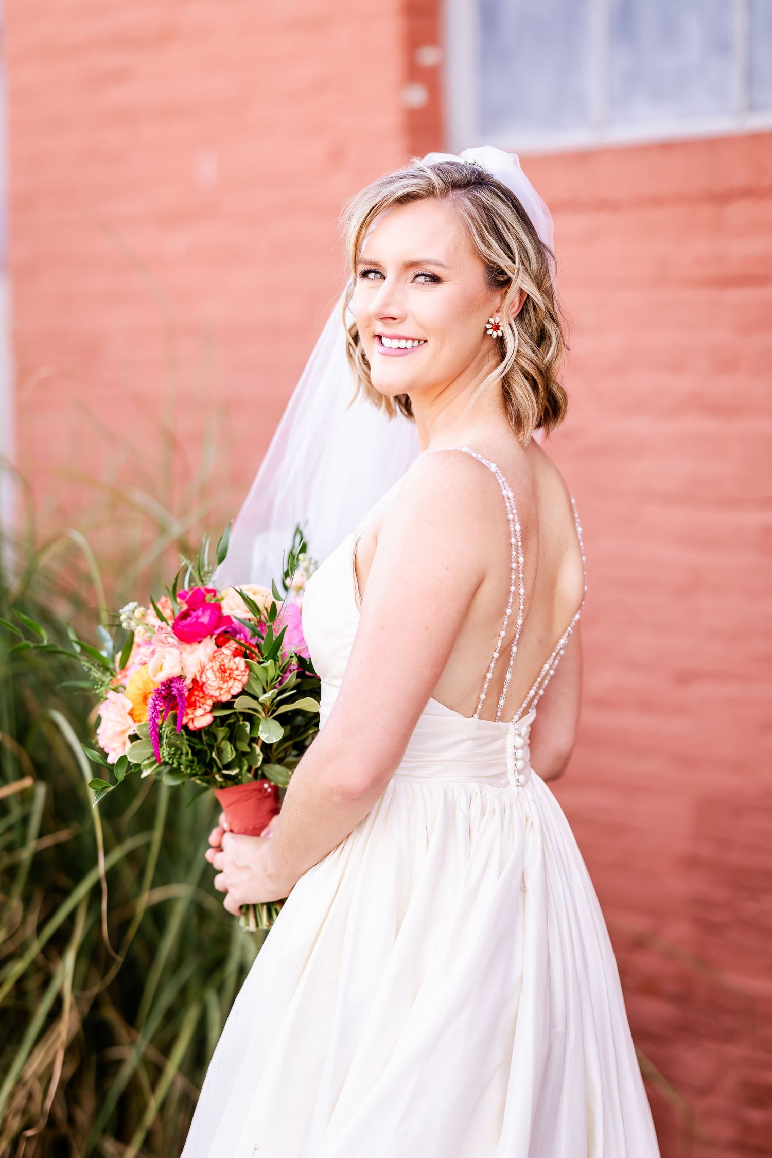 Bride in elegant white gown holds colorful bouquet while posing against coral-colored wall.