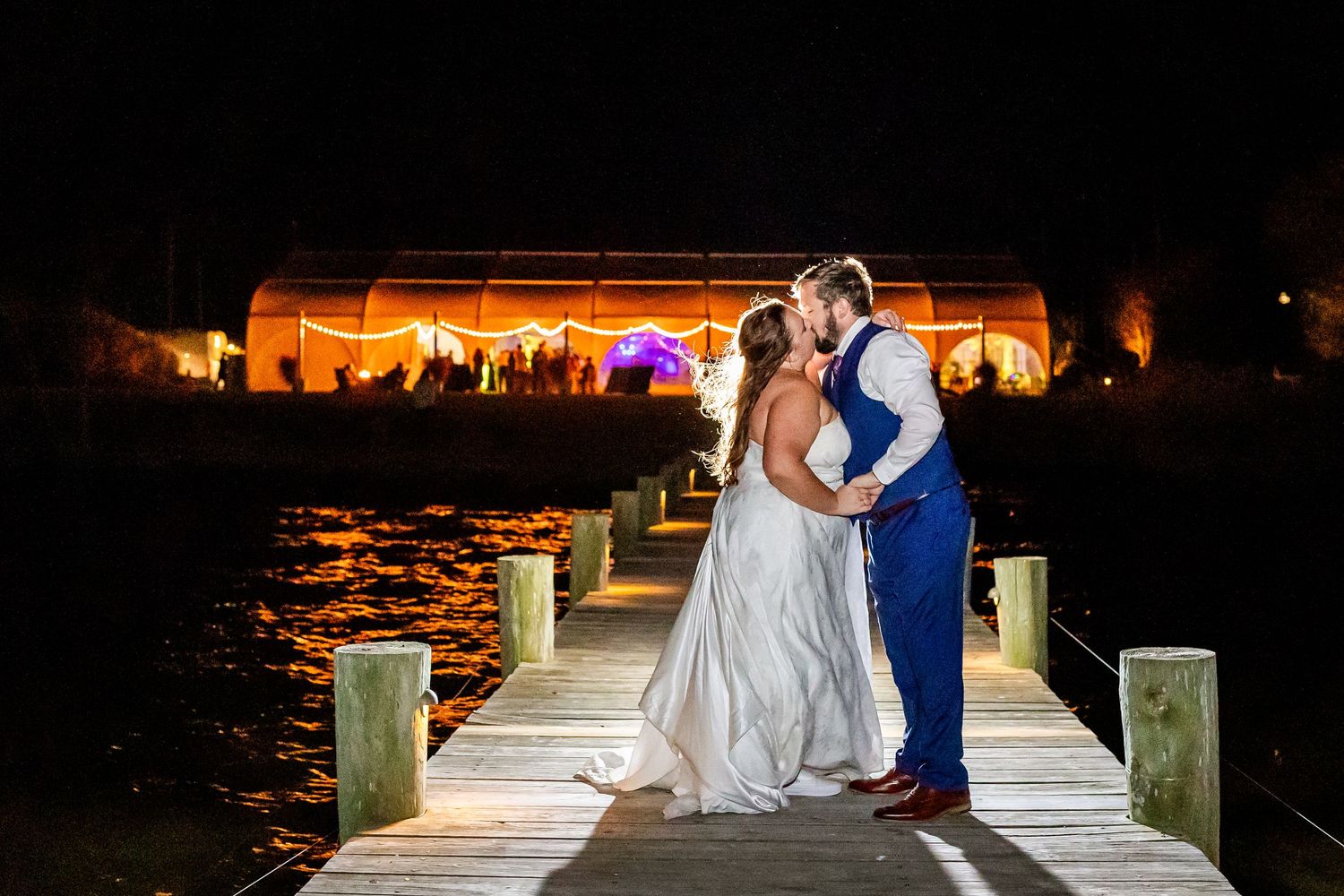 Nighttime dock wedding scene with illuminated venue in background and colorful lighting.