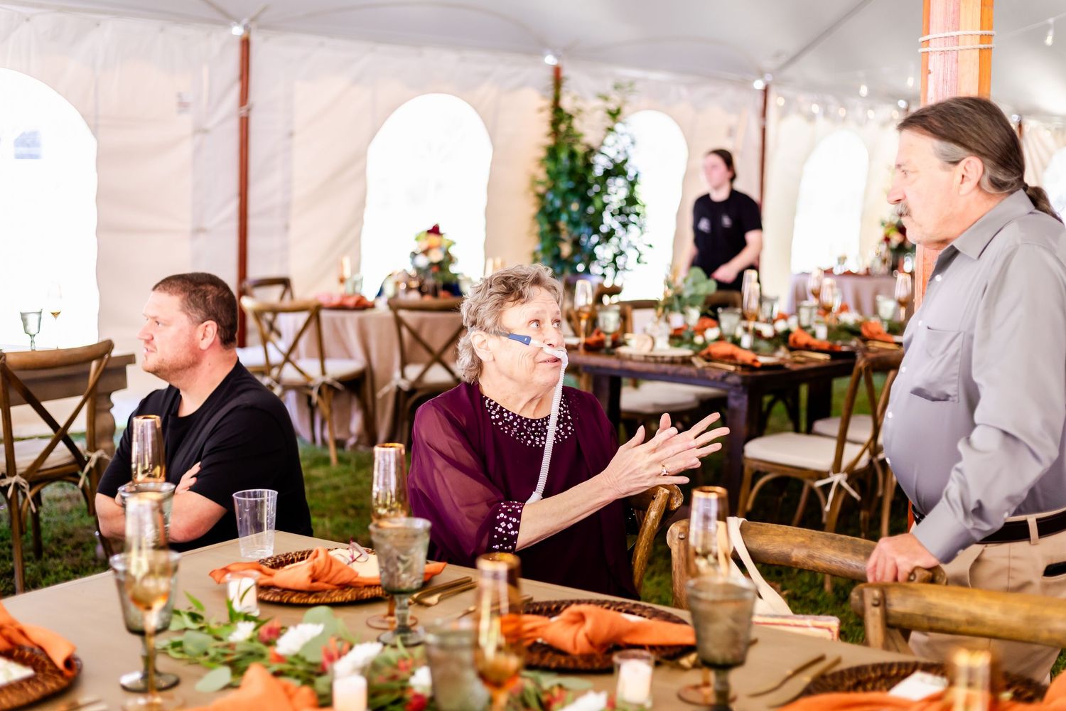Guests engage in lively conversation at a rustic wedding reception under a white tent.