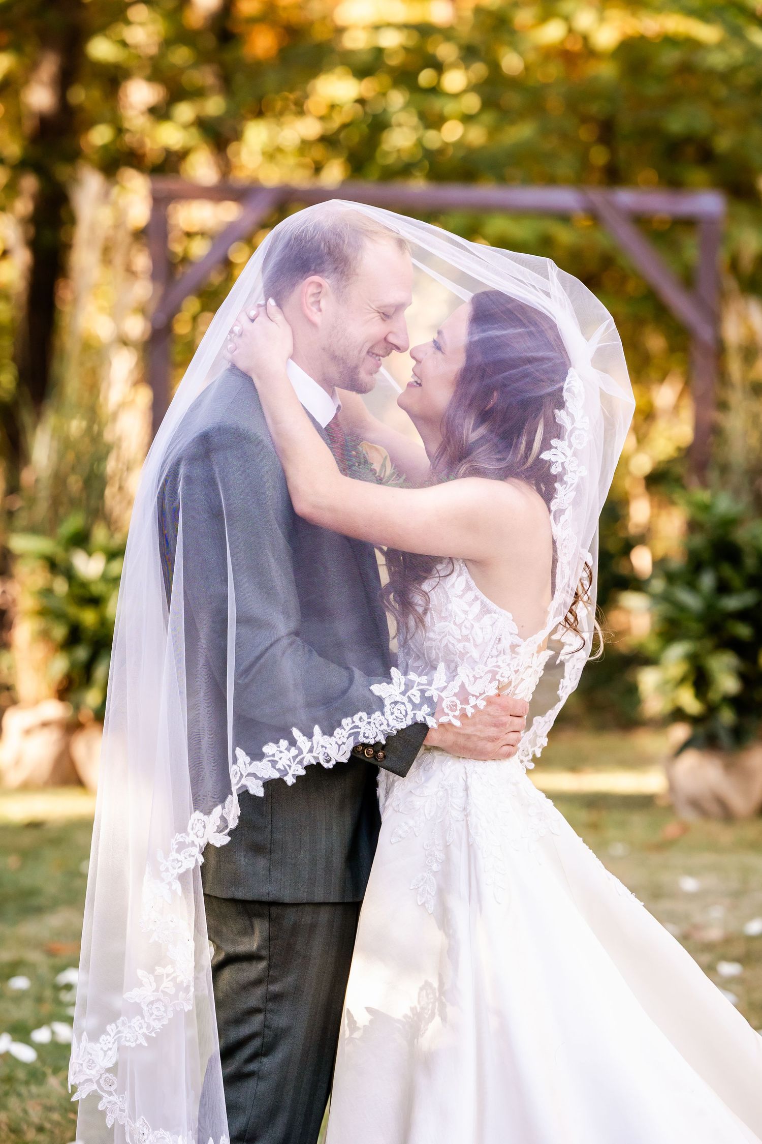 A romantic wedding couple embraces under a flowing veil in a sunlit outdoor setting.