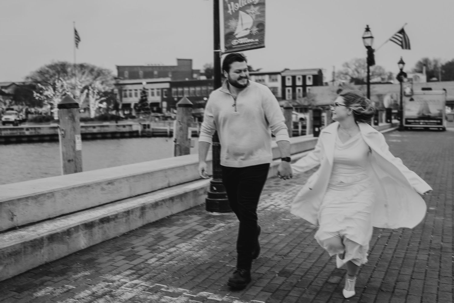 A couple walks and dances together on a brick path near a waterfront during what appears to be a wedding celebration.