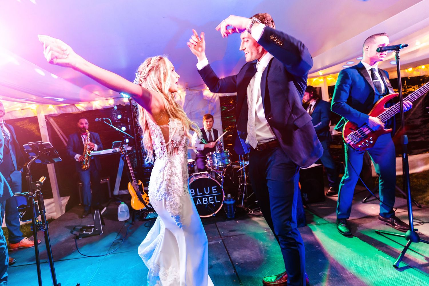 Newlyweds dancing energetically with live band performing on stage at wedding reception with colorful lighting effects.