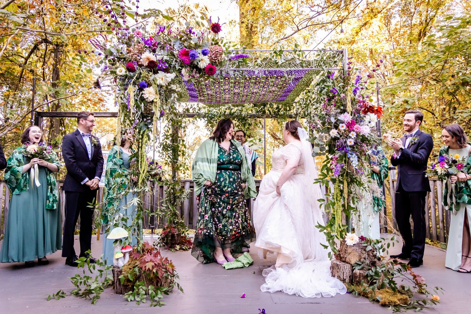 Outdoor wedding ceremony under a floral arch decorated with purple and pink blooms.