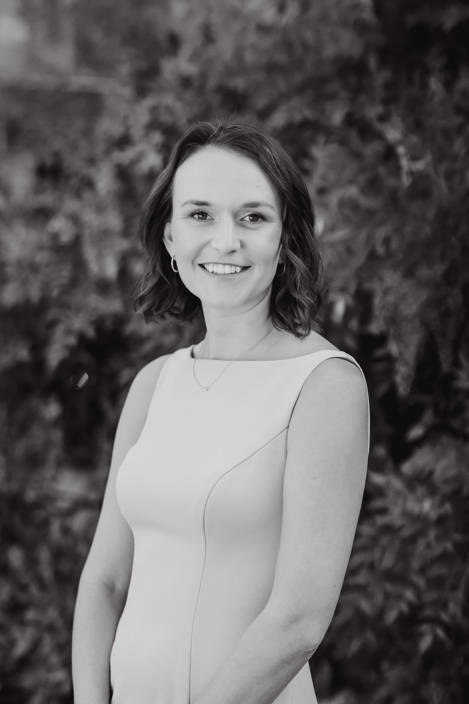 Professional black and white portrait of someone in a sleeveless white dress against a blurred natural background.