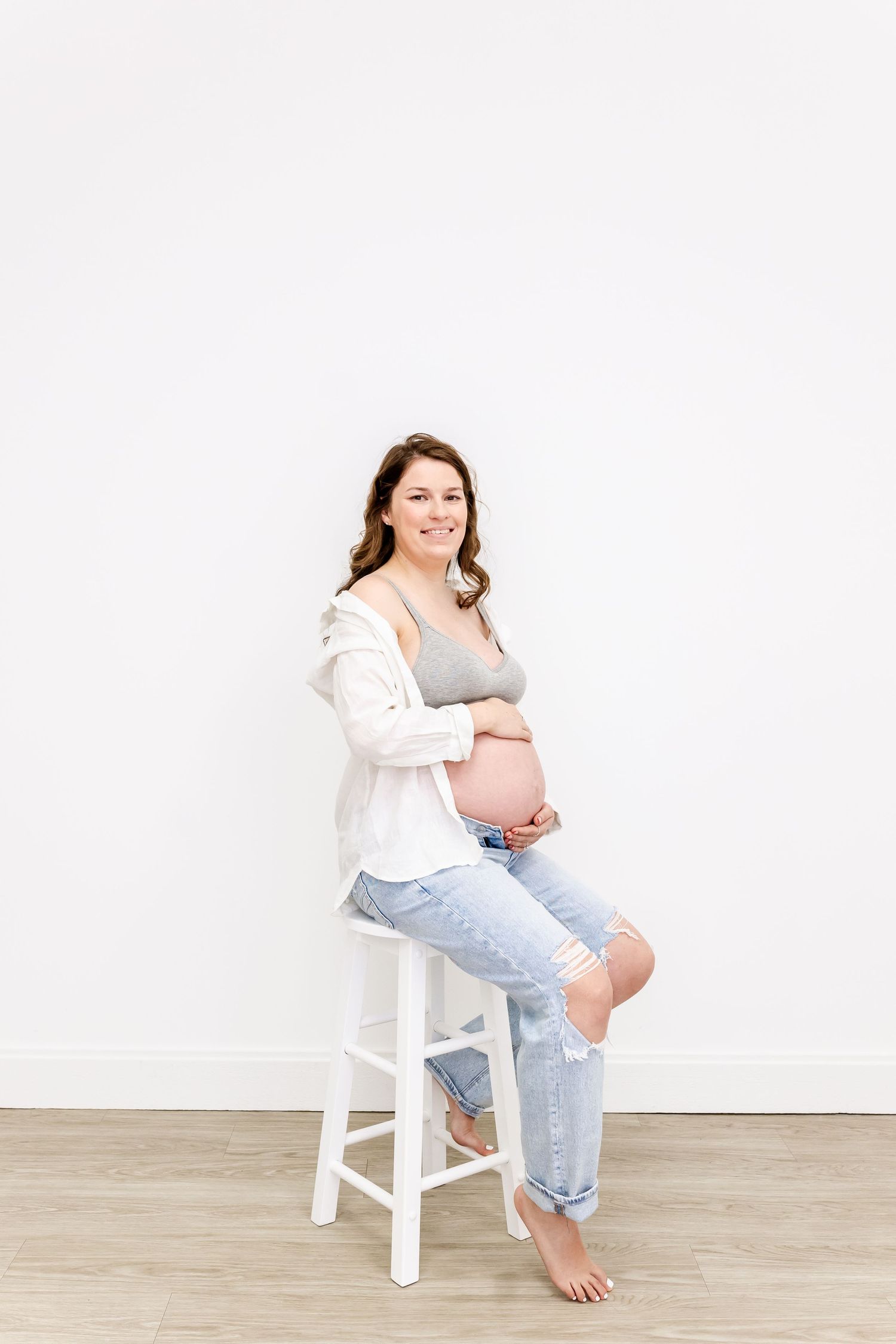 Pregnant woman in casual attire sitting on white stool against plain wall, smiling and cradling baby bump.