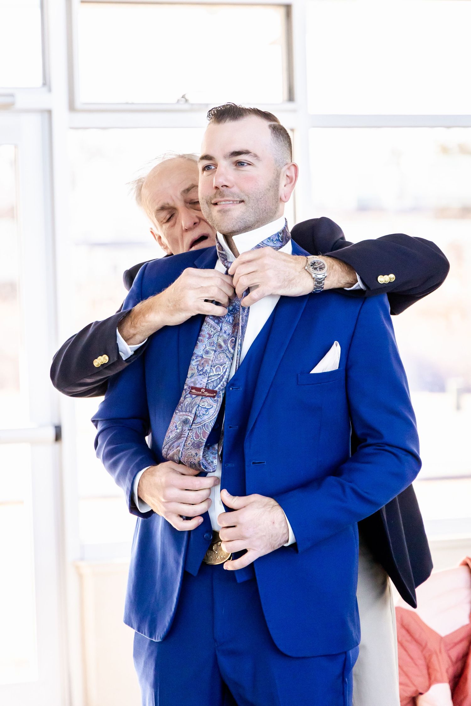 Two people in formalwear help finish getting dressed in a bright indoor setting with a blue suit being adjusted.