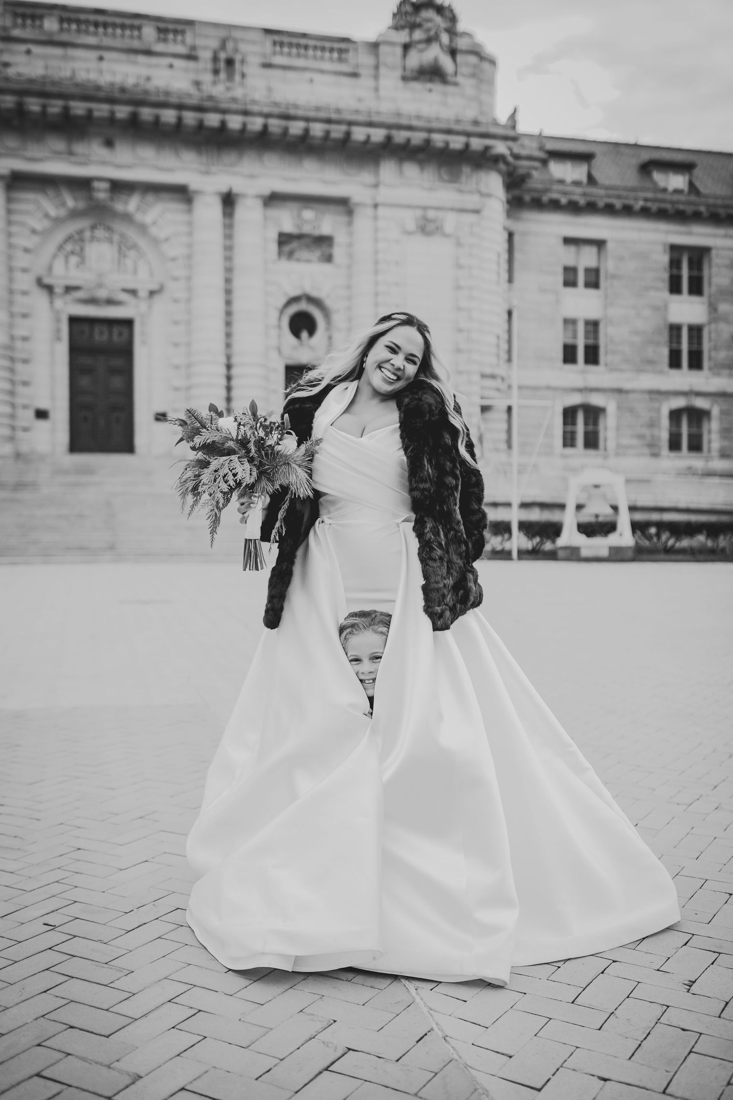 A bride in a flowing white gown holds a bouquet while standing outside a historic building in black and white photography.