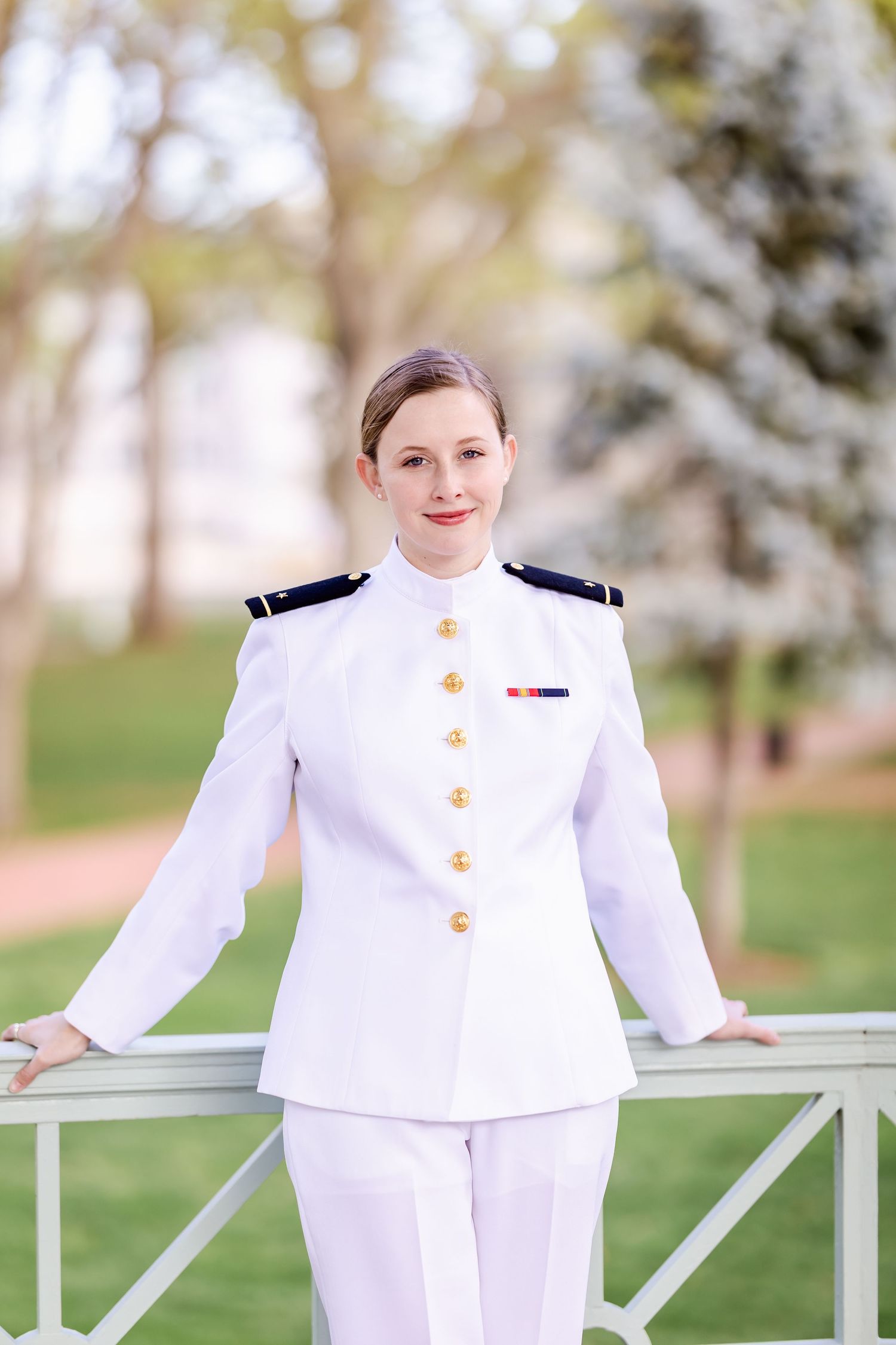 A service member in dress white naval uniform stands with hands on railing in a spring garden setting.
