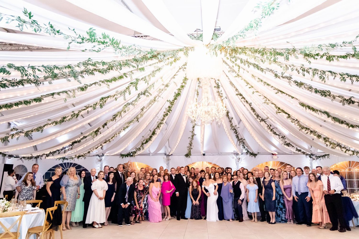 Large wedding reception under an elegant white tent canopy with draped fabric and greenery decorations.