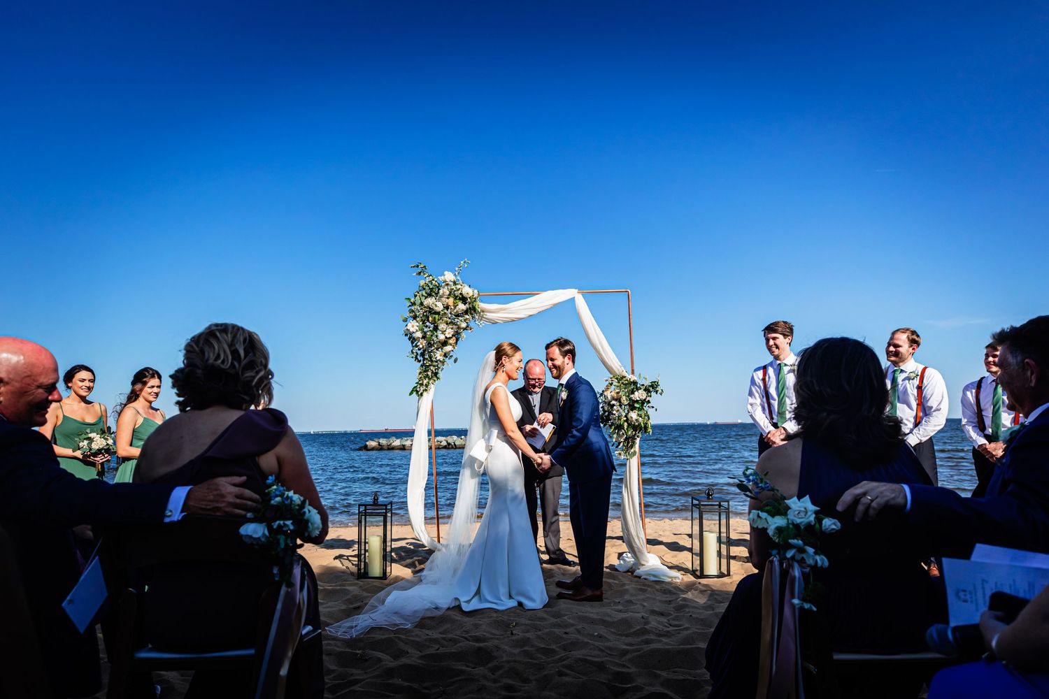 A beach wedding ceremony under a white arch with guests seated on either side against a bright blue ocean backdrop.