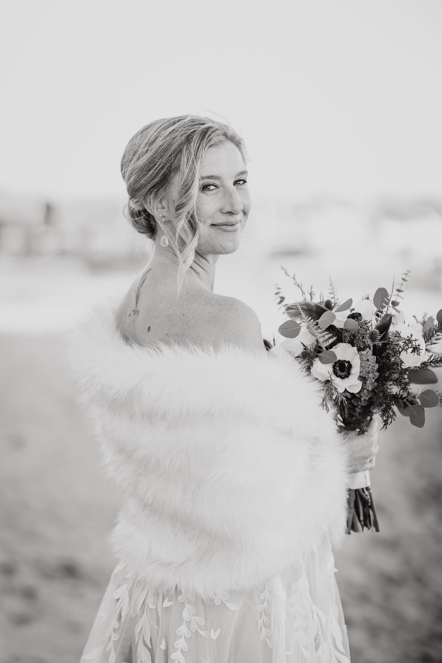Black and white wedding portrait showing bride in white dress with fur wrap holding floral bouquet outdoors.