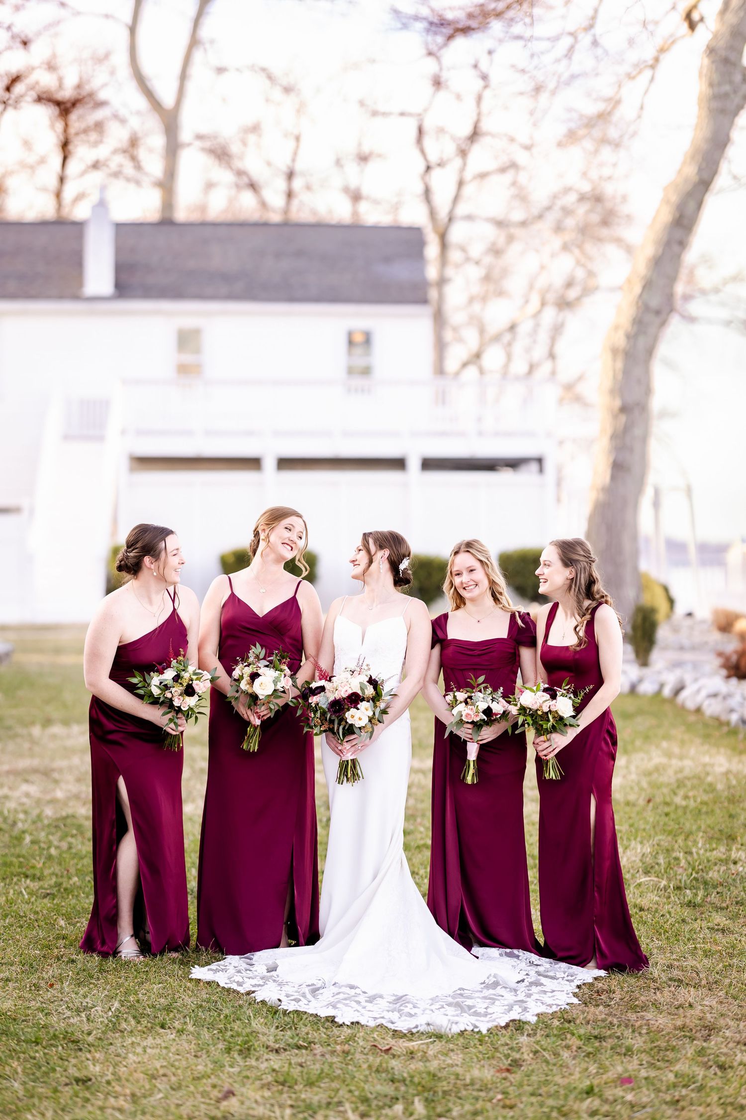 A bridal party in burgundy bridesmaid dresses and white wedding gown pose together outdoors on a grassy lawn.
