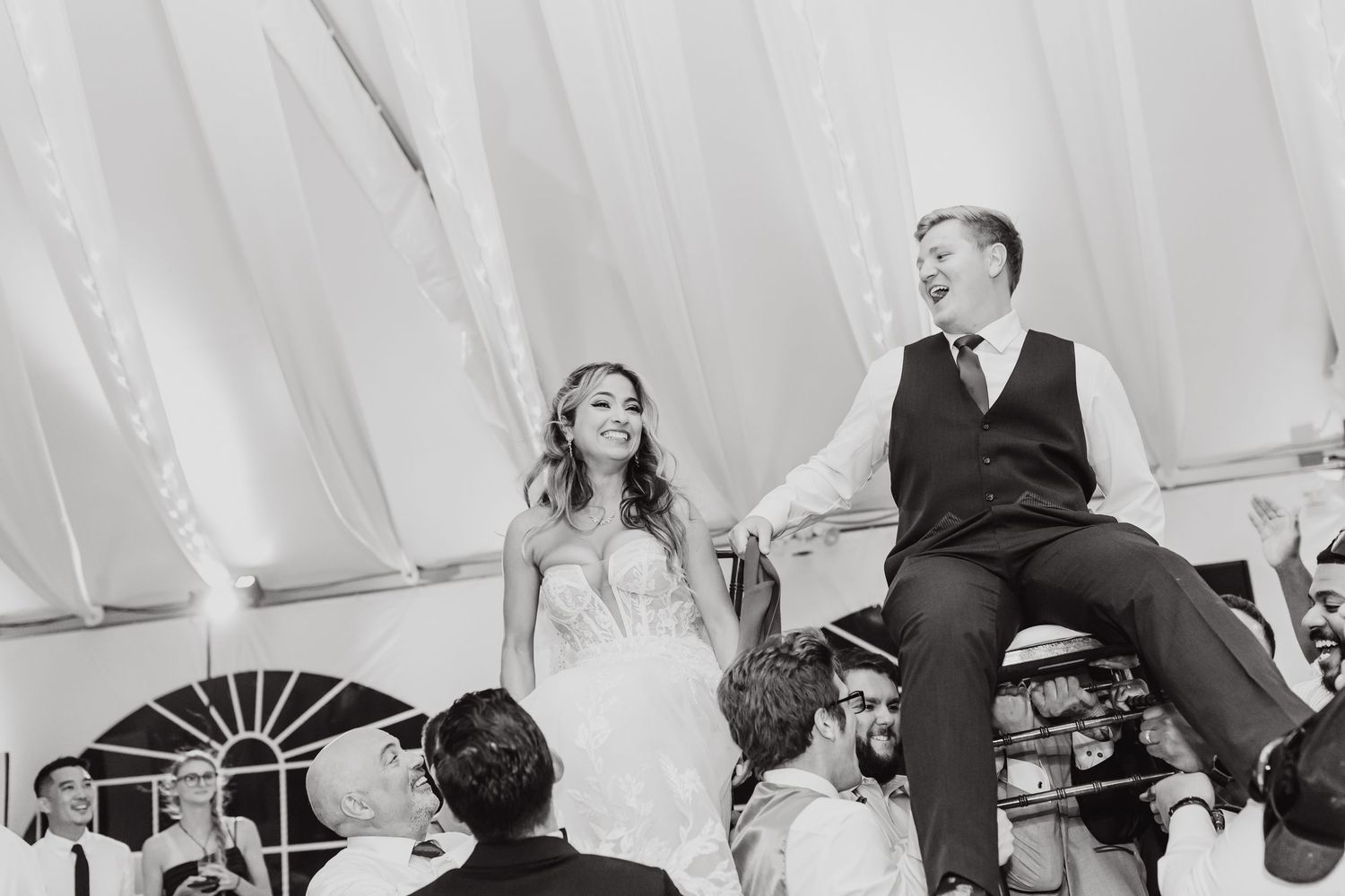 Wedding celebration shows guests lifting the couple in chairs during a traditional hora dance at an elegant tent reception.