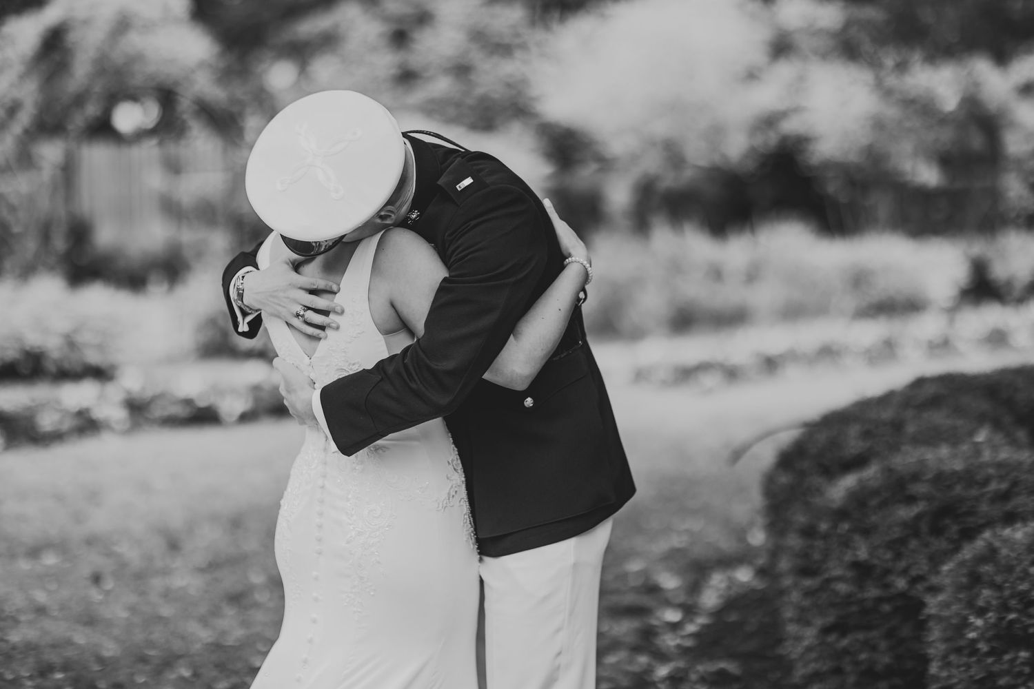 A romantic black and white photo of a couple embracing outdoors, with the bride wearing a white dress and groom in dark suit.