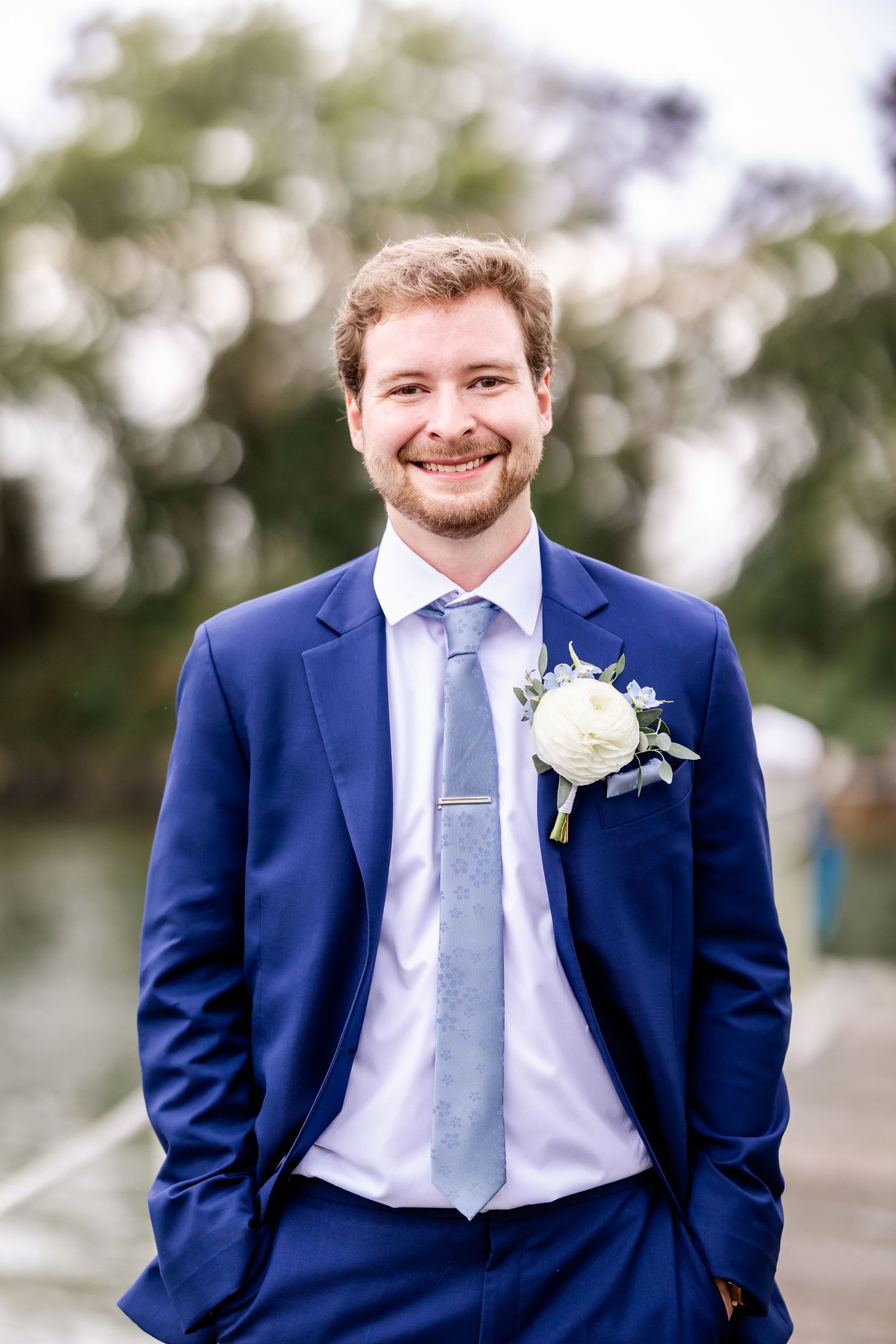 Groom in navy suit and light blue tie smiles at camera in outdoor portrait series.