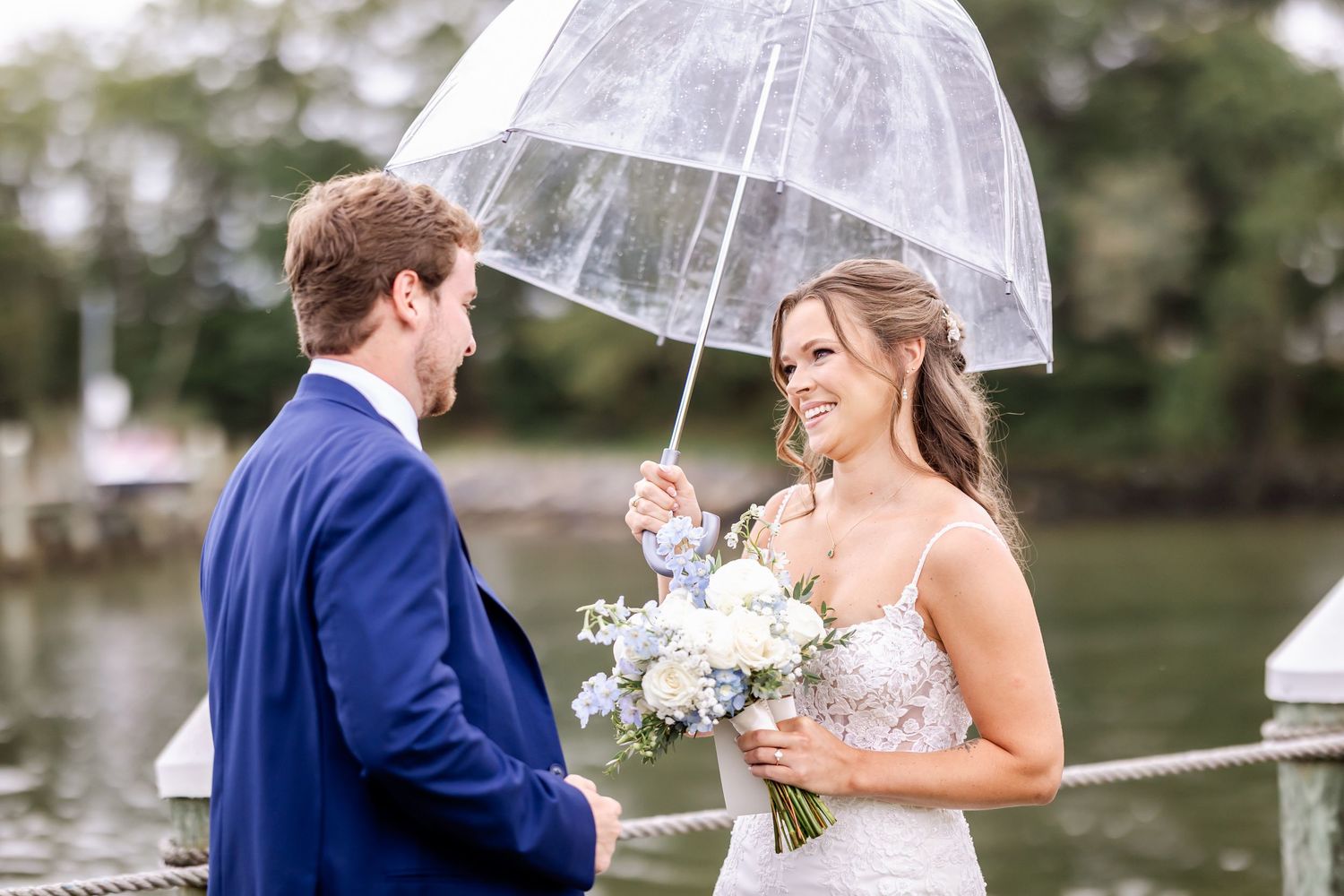 Wedding couple shares romantic moments under a clear umbrella by the water.