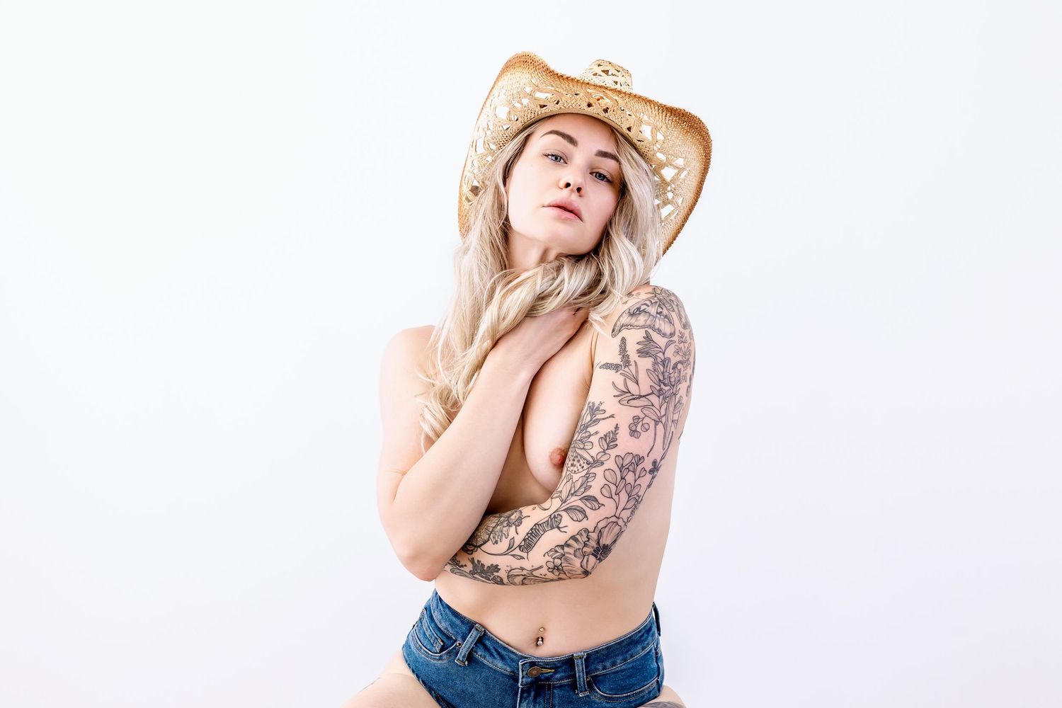 Topless woman in cowboy hat and denim shorts poses against white background with floral arm tattoo.