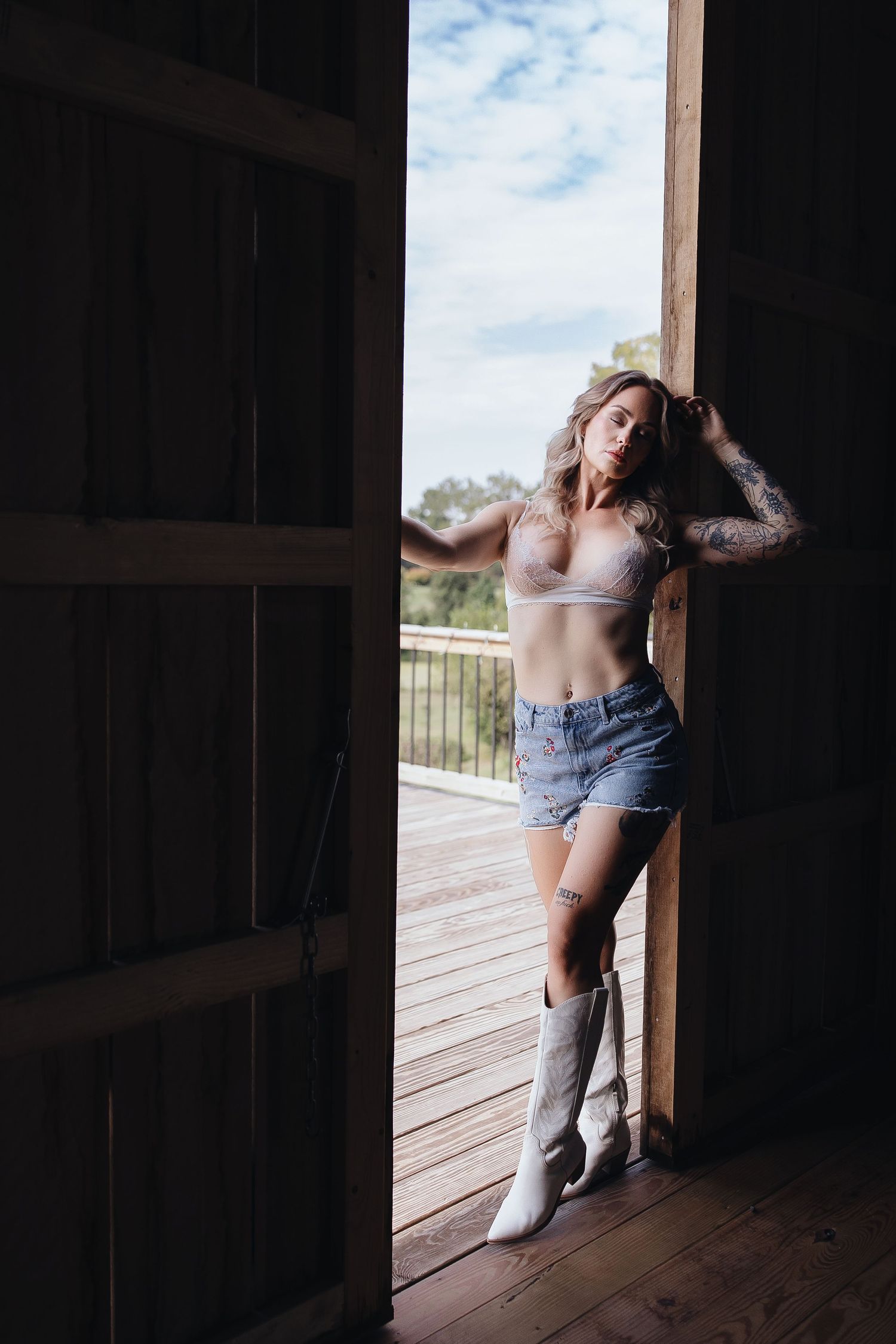 Woman in denim shorts, crop top, and white cowboy boots posing in rustic barn doorway with blue sky backdrop.