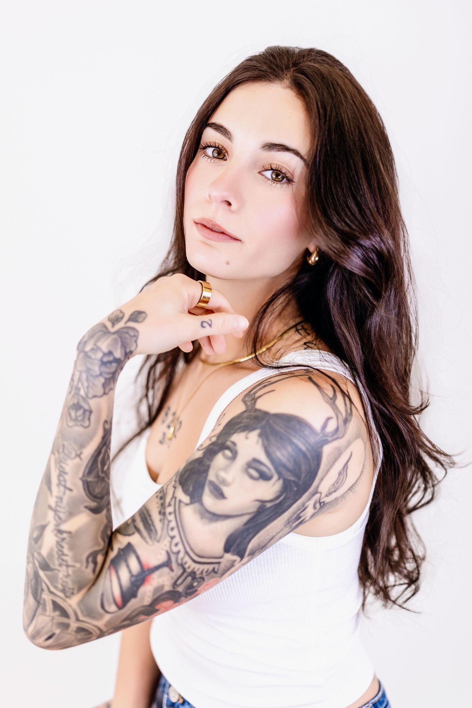 Young woman with long dark hair and full sleeve tattoos poses in white tank top against white background.