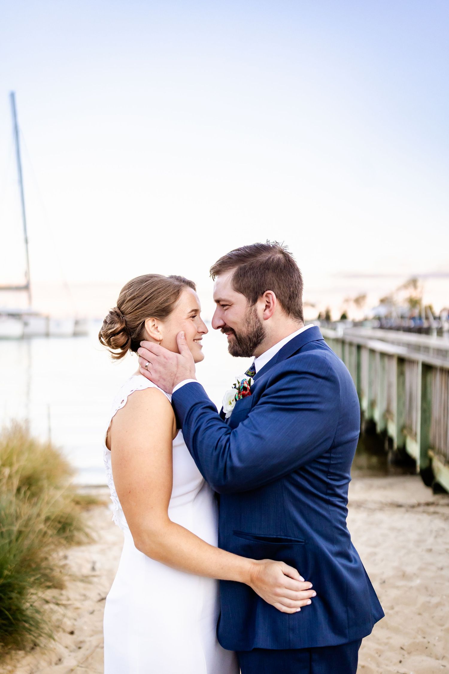 A bride and groom share a tender moment on a sandy beach near a wooden pier on their wedding day.