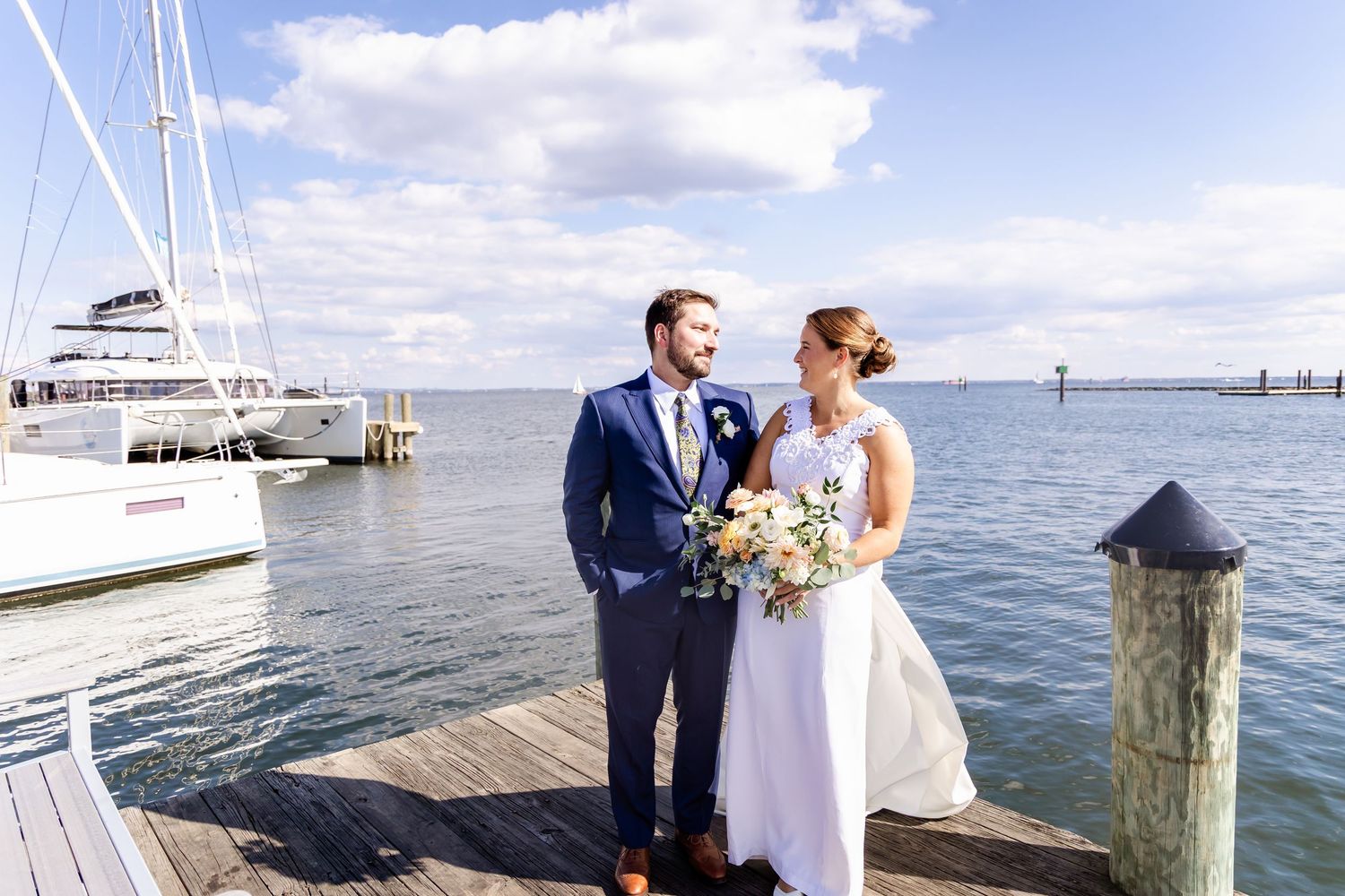 Bride and groom share a romantic moment on a waterfront dock with sailboats and blue sky behind them.