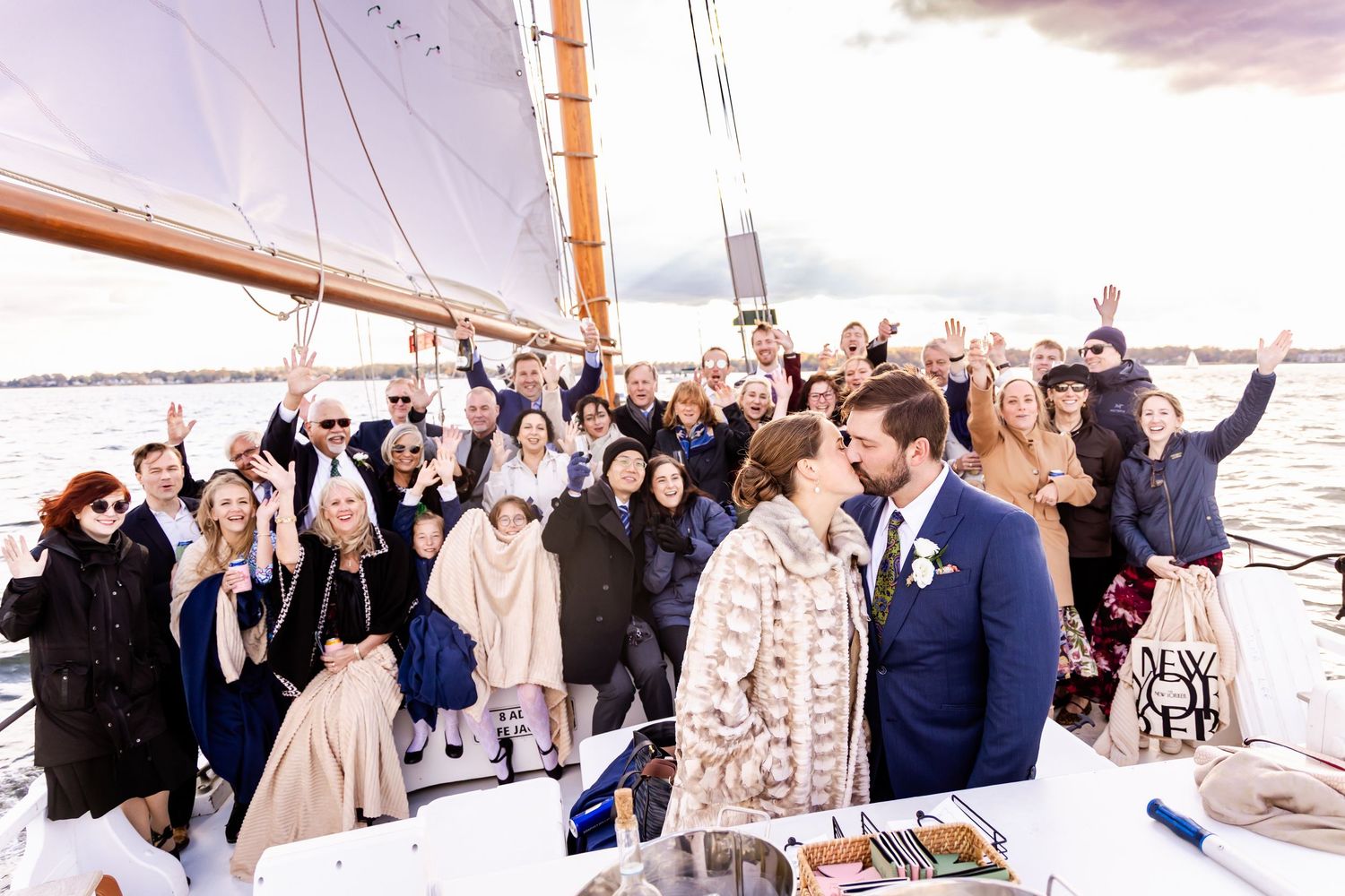 Wedding party celebrating on a sailboat deck, with bride and groom kissing surrounded by cheering guests on open water.