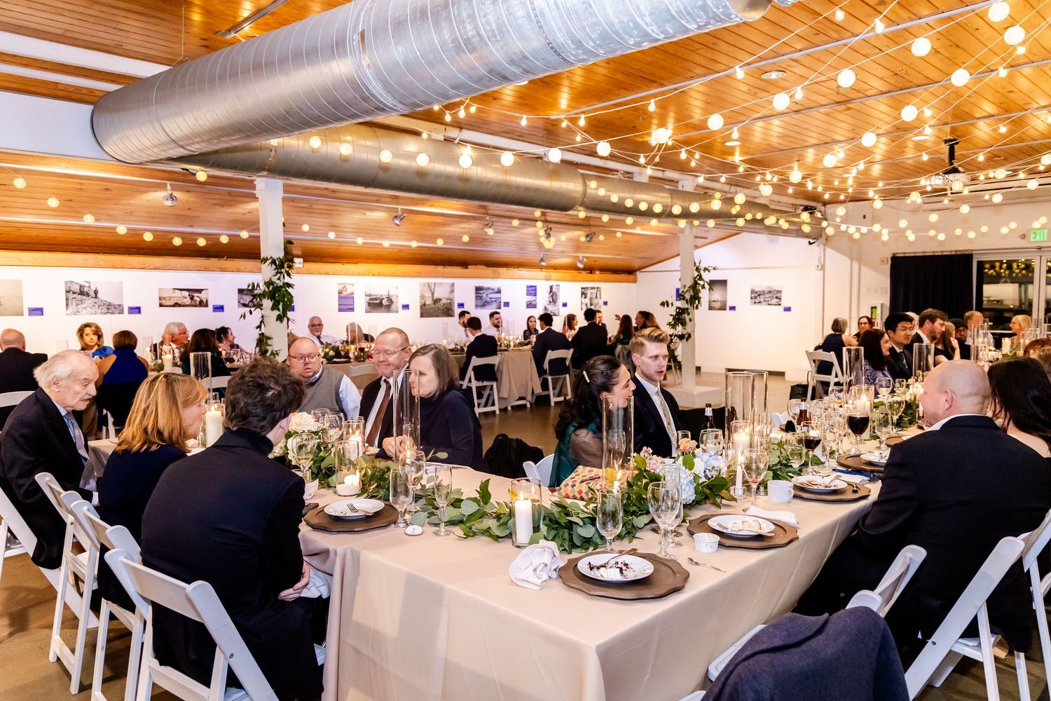 Elegant wedding reception in a modern venue with string lights, white chairs, and guests seated at decorated tables.