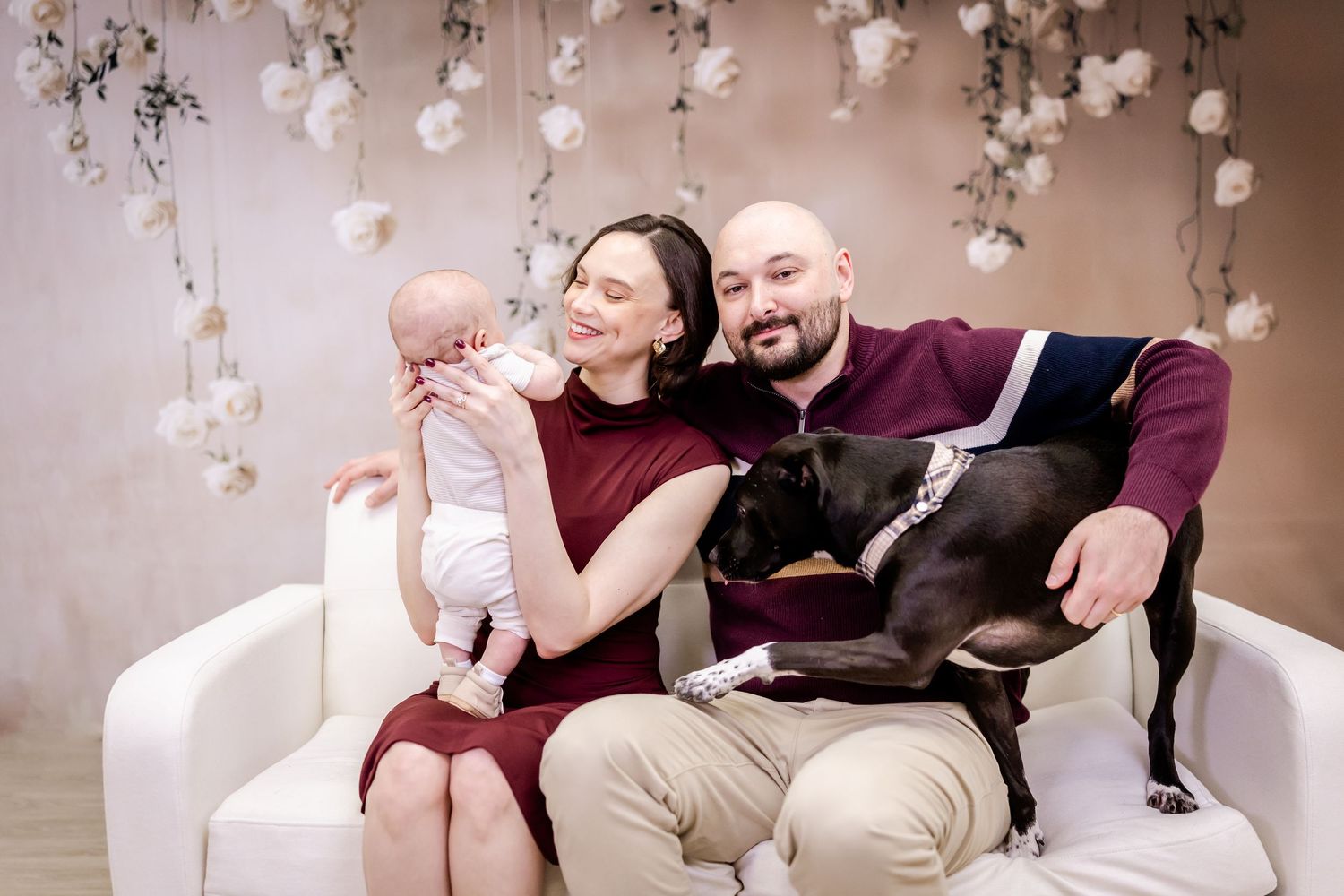 A family sits together on a white couch with their black dog under hanging floral decorations.