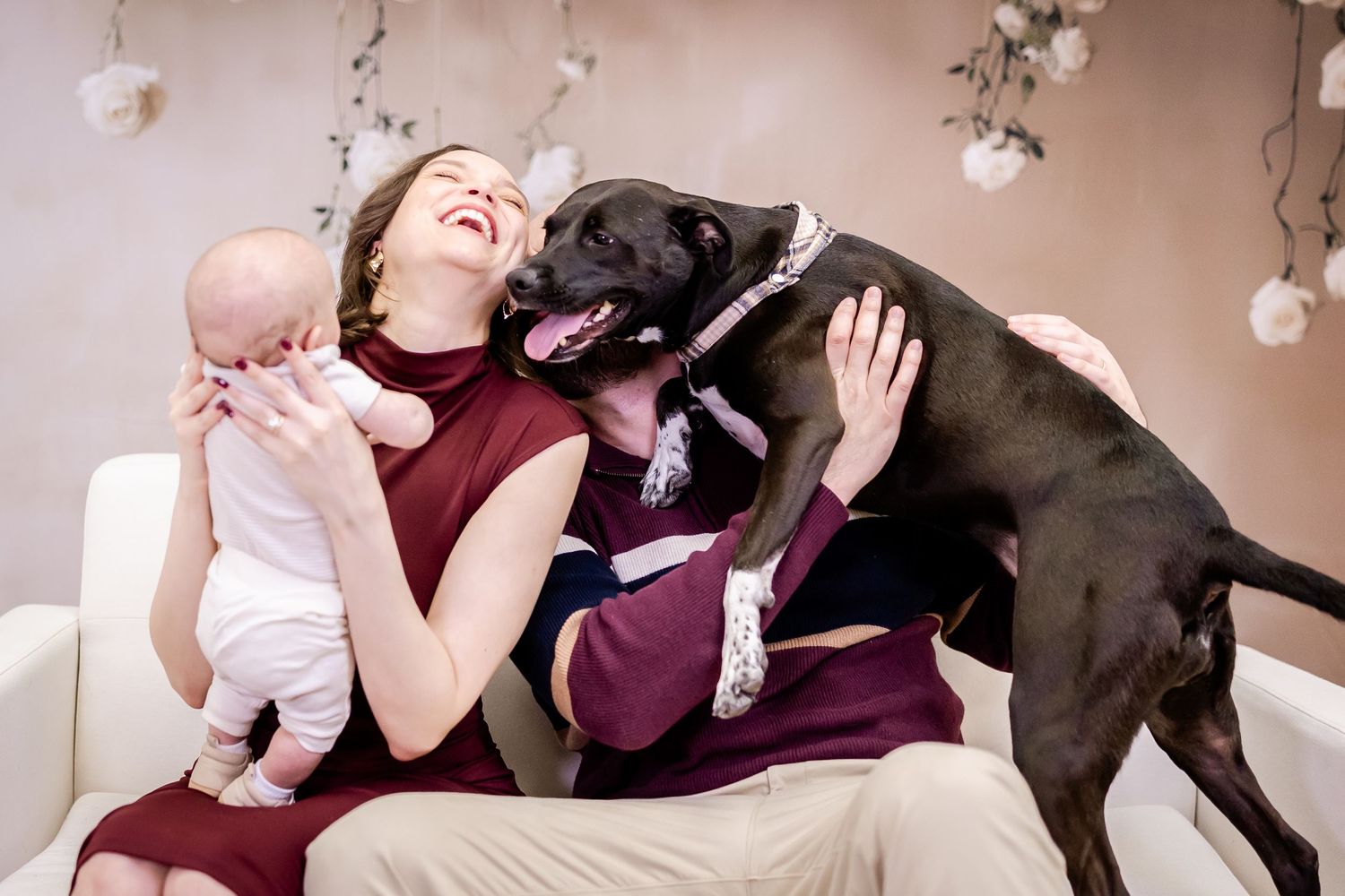 A joyful family moment with a black dog giving kisses while sitting together on a couch with floral decorations.