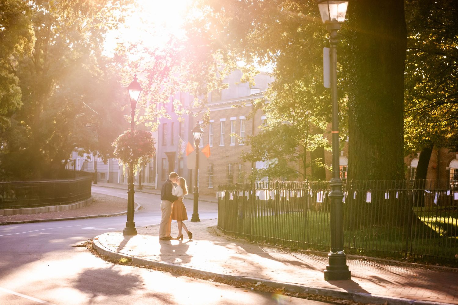 Sunlit urban park pathway with iron fence and street lamps casting warm golden light during sunset.