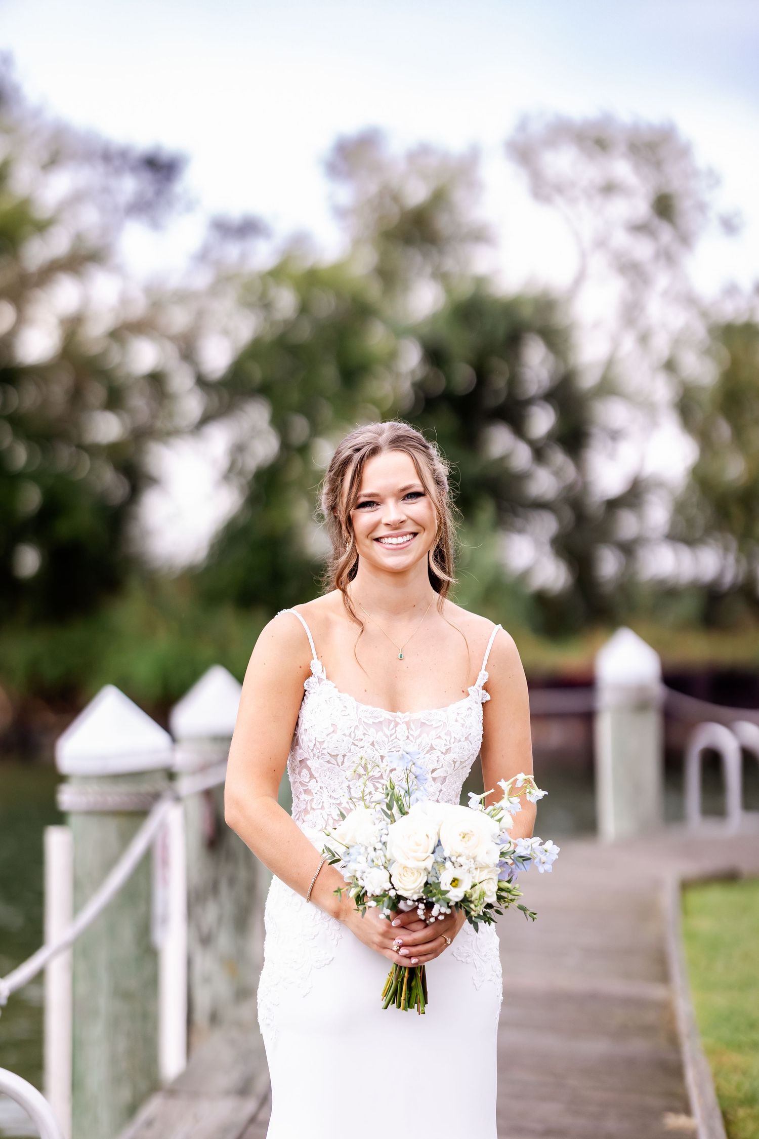 A bride in a white lace wedding dress stands on a wooden dock holding a bouquet of white flowers on her wedding day.