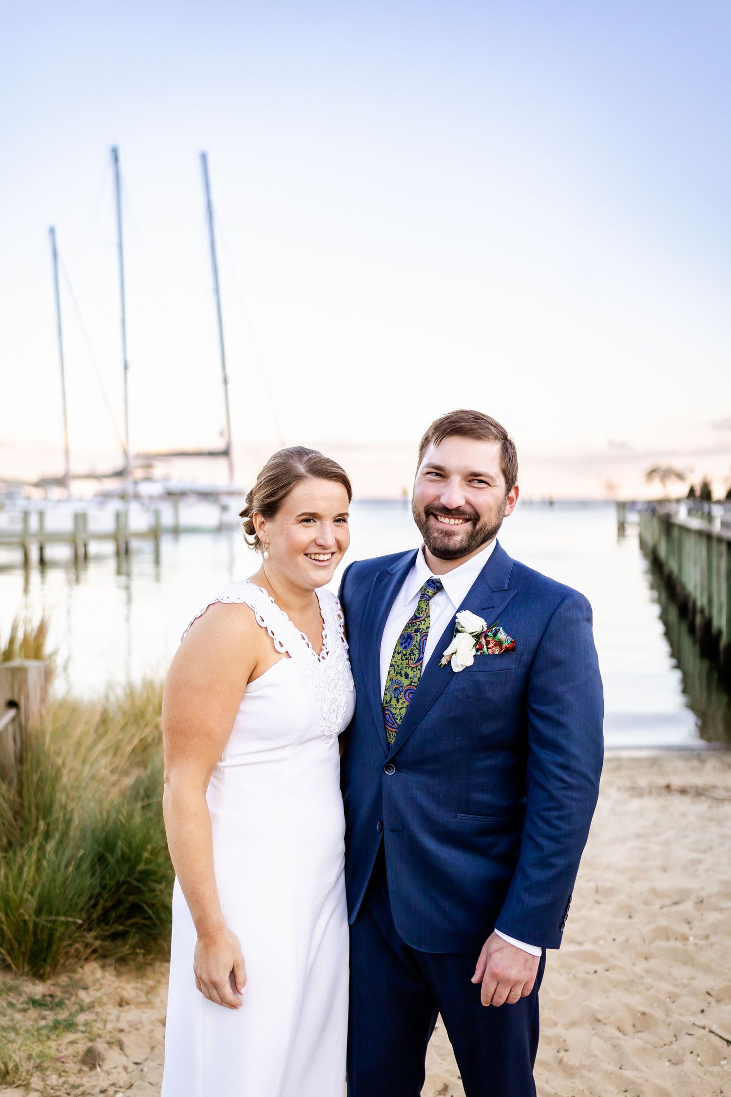 Smiling bride in white dress and groom in navy suit pose together on sandy waterfront at sunset.