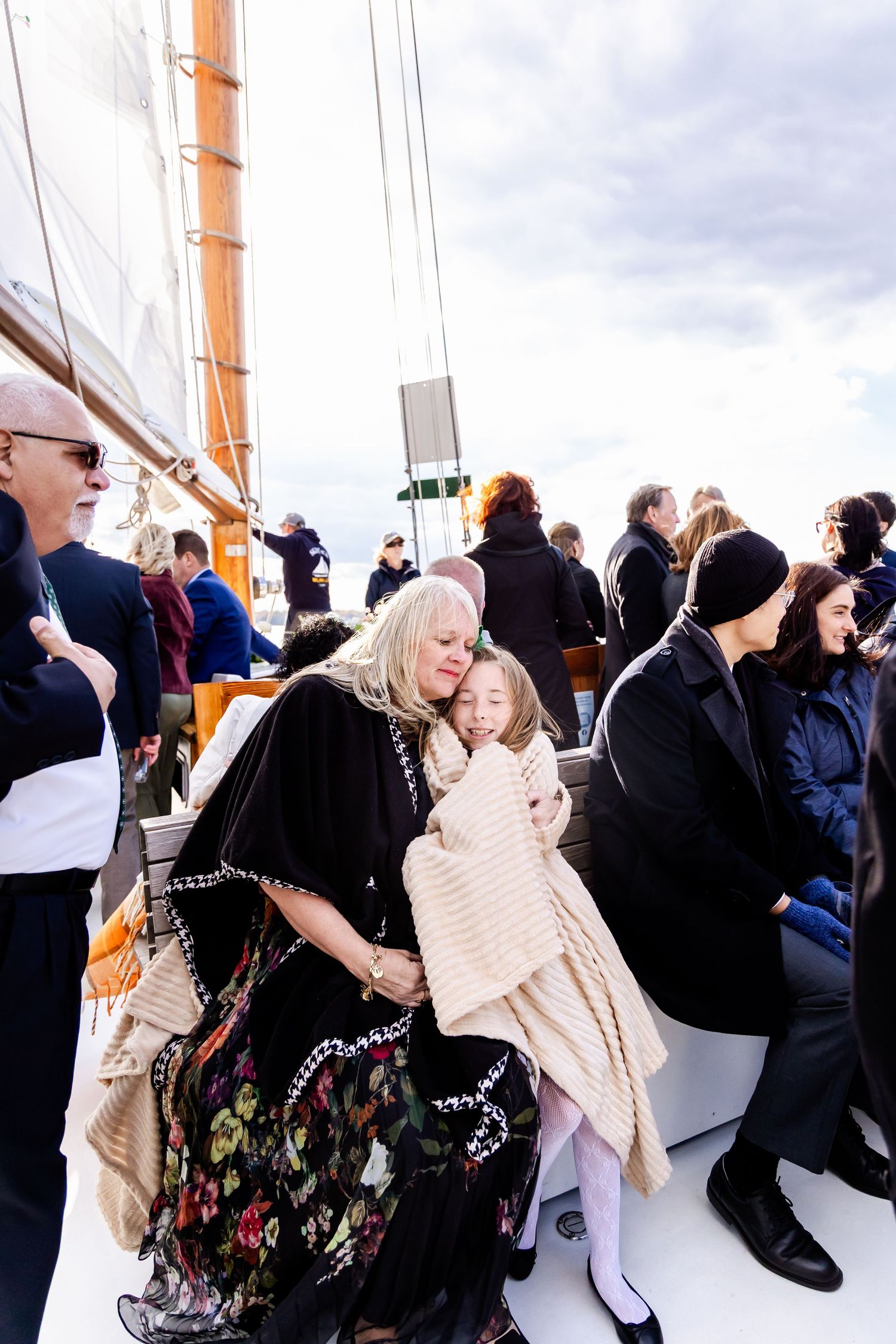 Grandmother hugging granddaughter wrapped in blanket on a crowded sailboat deck under cloudy skies.