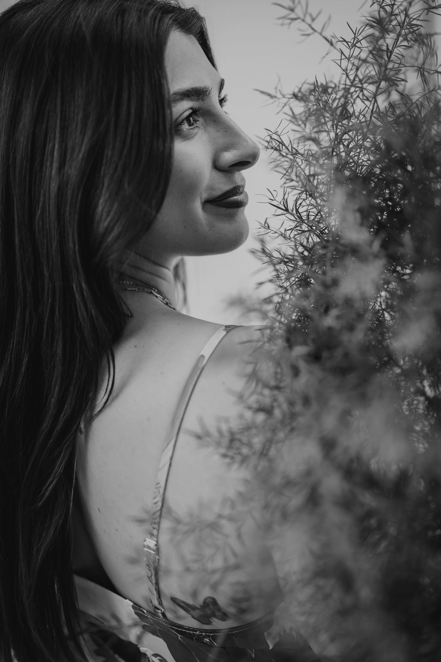 Black and white portrait of a young woman with long dark hair beside lush green foliage, gazing thoughtfully.