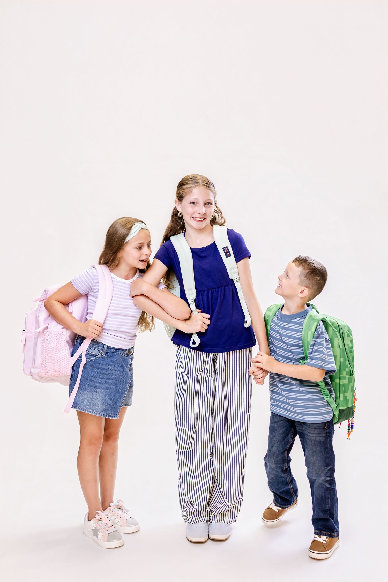 Three school children with backpacks smile and hug together against a white background.