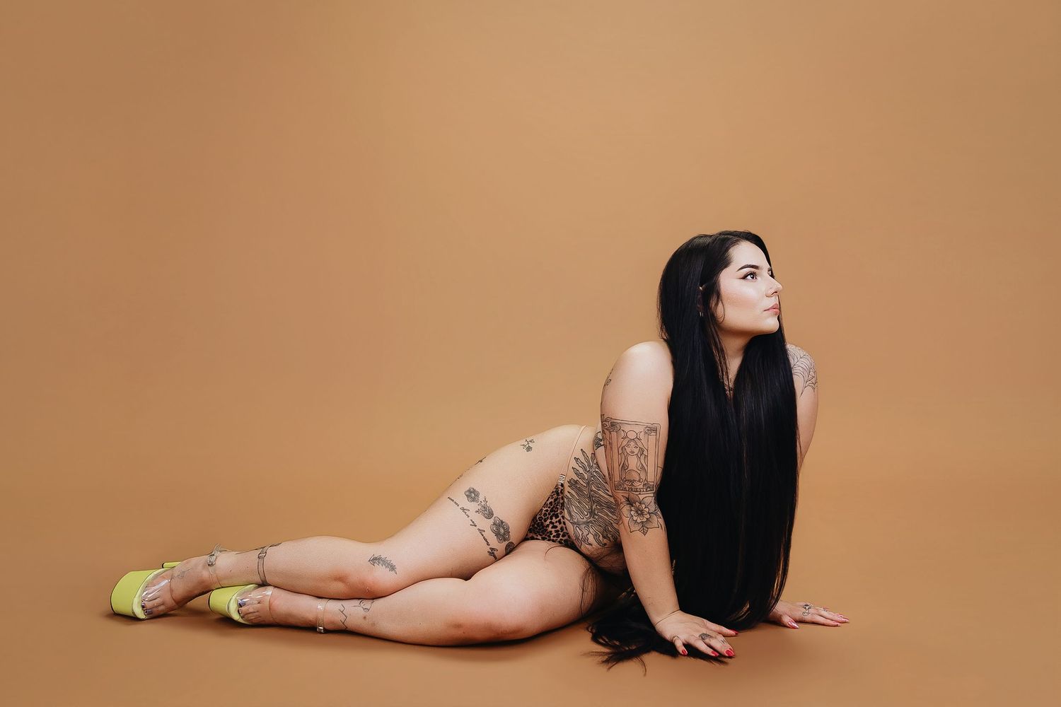 Tattooed woman with long black hair posing on floor in nude bodysuit and yellow heels against tan background.