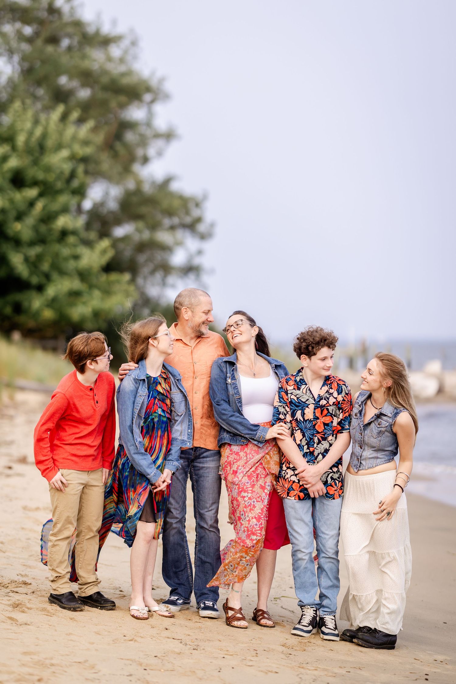 A family of six poses playfully on a sandy beach, wearing coordinated casual outfits in blue and red tones.