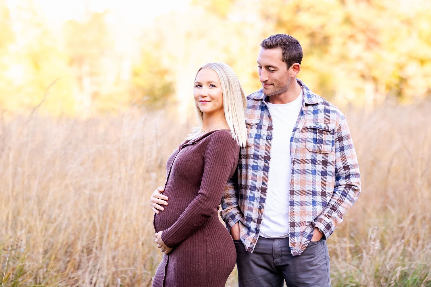 A couple stands together in a golden field during sunset wearing plaid and brown casual clothing.