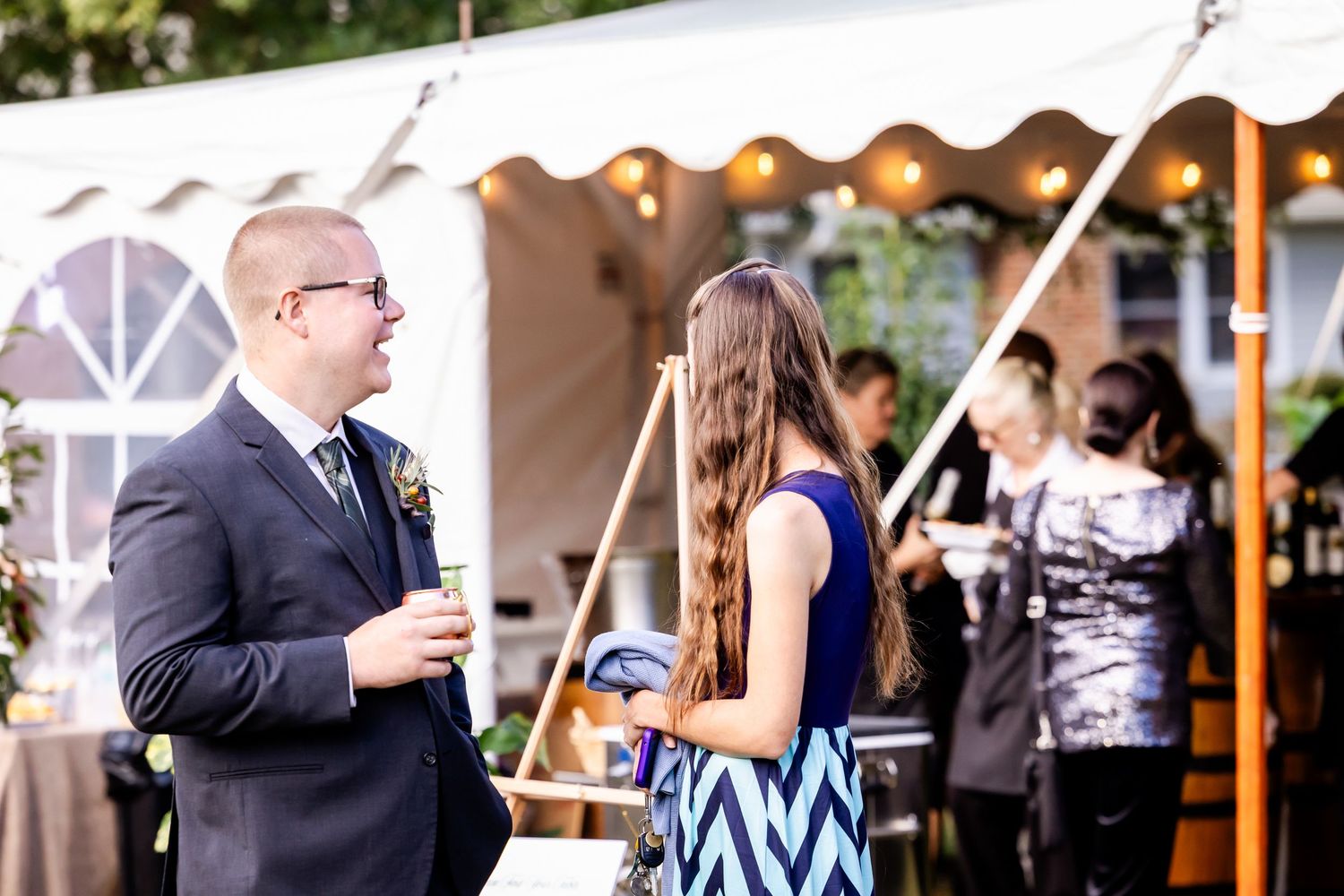Wedding guests socializing at an outdoor reception under white tent with string lights.