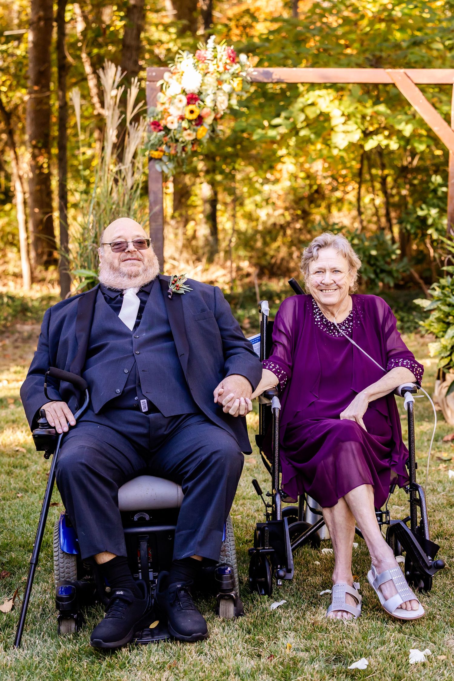 An elderly couple in formal attire holding hands while seated in wheelchairs under a floral wedding arch.