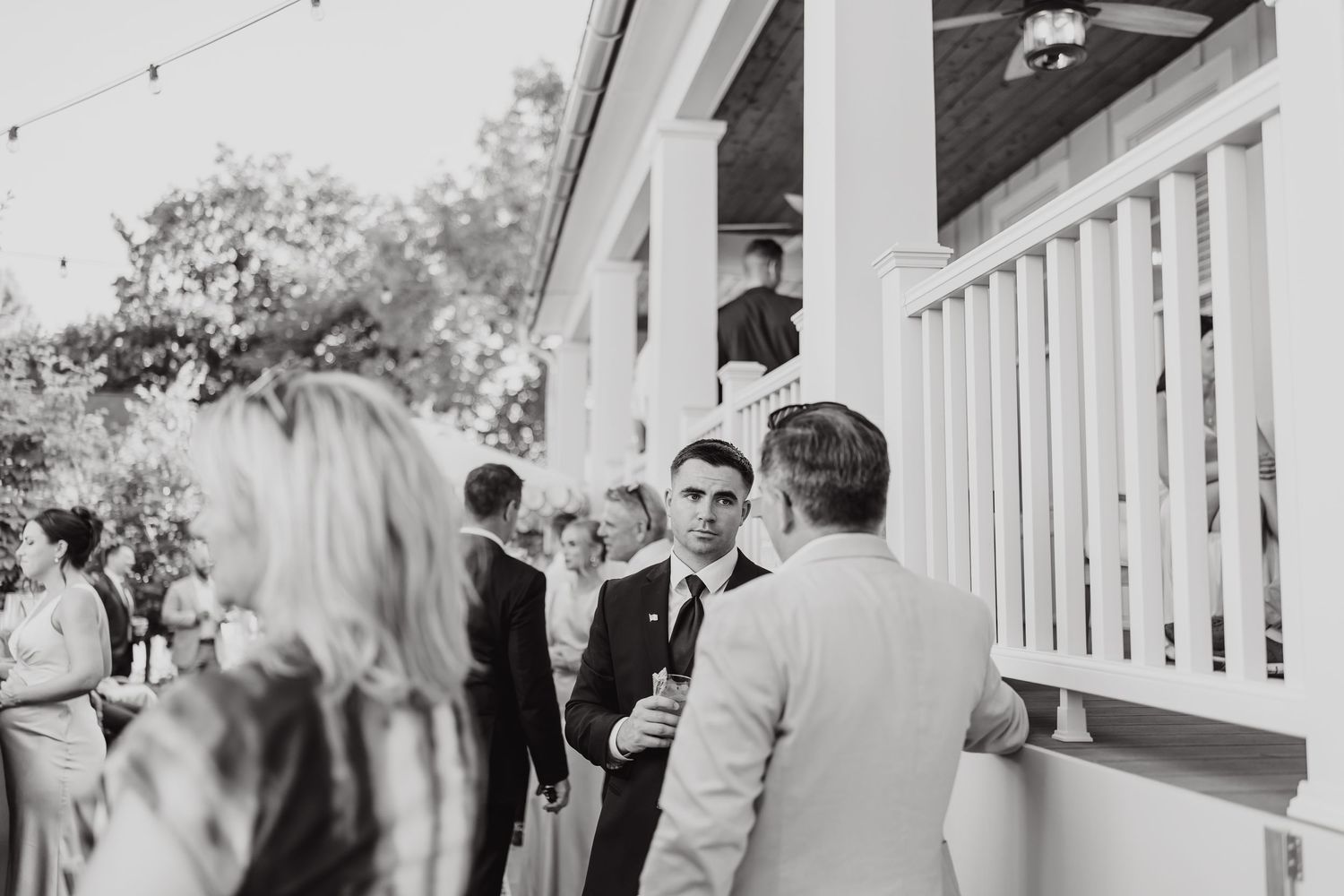 A black and white photograph showing wedding guests gathered on an outdoor porch with white railings.